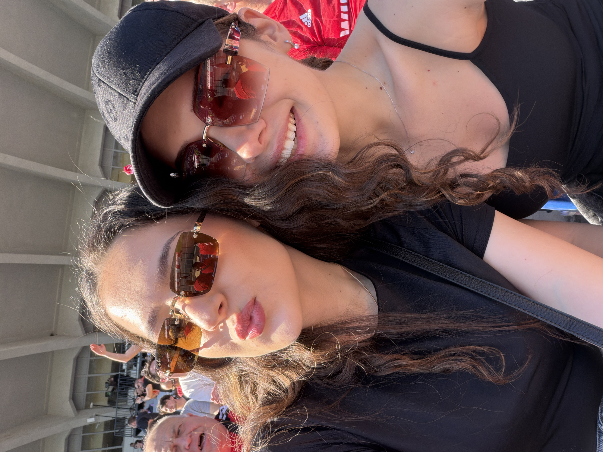 Two women are posing for a selfie in a crowded outdoor stadium. The woman on top is wearing a black baseball cap and sunglasses and has long, curly brown hair. She is smiling. The woman below is also wearing sunglasses and a black top. They are likely at a sporting event based on the crowd and red Adidas jersey visible in the background. The location may be Vasil Levski National Stadium in Sofia, Bulgaria, given the style of the stadium and presence of Adidas apparel often associated with Bulgarian sports teams.