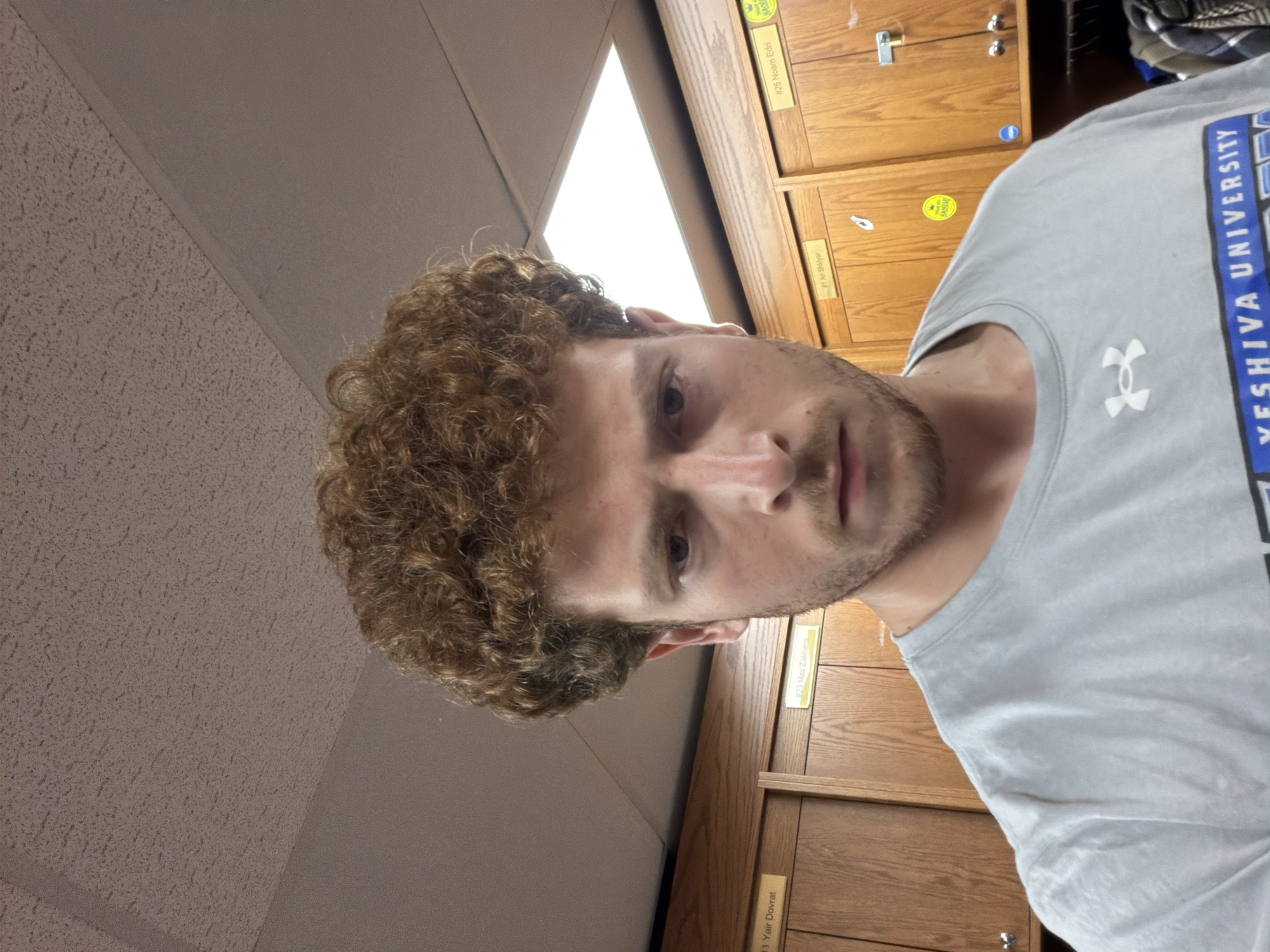 This is a tilted, close-up indoor portrait featuring a young man with curly, reddish-brown hair and a light beard, looking directly at the camera. He is wearing a light grey t-shirt with "YESHIVA UNIVERSITY" printed vertically in blue on the right side and a white Under Armour logo on the chest.
The background reveals a series of light brown wooden lockers or cabinets, some featuring small white and yellow labels. Visible text on these labels includes: "#25 Noam Edri", "#1 Ari Shklyar", "#23 Max Zakheim", and "#1 Yair Dovrat". A small yellow circular sticker with a crown and text (likely "Happy Birthday") is present, along with a partially visible label reading "HASH". Above the lockers, a bright fluorescent light fixture illuminates an off-white ceiling composed of acoustic tiles.
The setting appears to be an institutional environment like a school or university, which aligns with the "YESHIVA UNIVERSITY" text on the shirt and the given location context of New York, United States of America. The lighting is bright and artificial, suggesting an indoor daytime or evening setting.