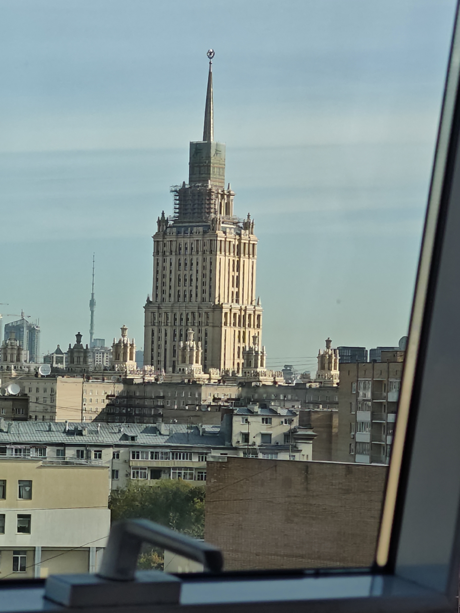The image captures a distant view of the Stalinist skyscraper, the Ministry of Foreign Affairs Building, located in Moscow, Russia. The sun illuminates the building, highlighting its intricate facade and spire topped with a star. Construction scaffolding is visible around the upper sections of the tower, suggesting ongoing renovation or maintenance. In the background, other buildings and a slender radio tower punctuate the Moscow skyline under a clear, pale blue sky. The foreground features a window frame and a metal handle, framing the cityscape.