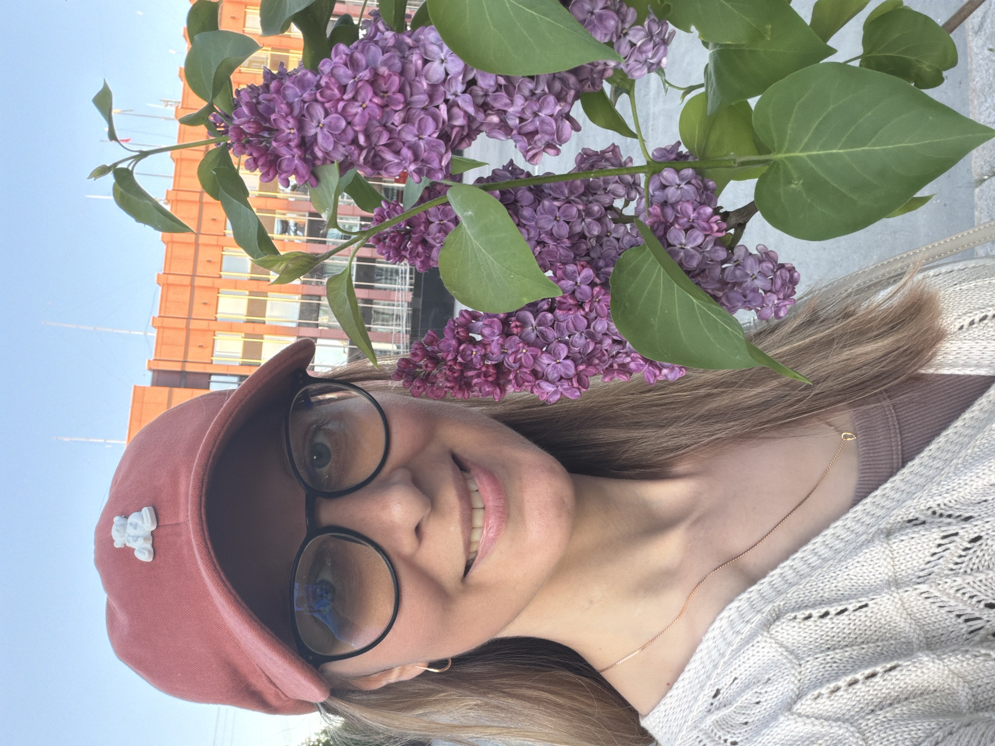 A photograph depicts a fair-skinned woman wearing a pink cap with a teddy bear emblem and circular black eyeglasses, smiling directly at the camera. She has light brown hair and is wearing a light-colored knit sweater with a gold chain necklace. A bouquet of purple lilacs is positioned in the foreground. The background reveals an orange building and a patch of blue sky, suggesting an outdoor setting. Based on the building's architecture, the location appears to be Izhevsk, Russia.