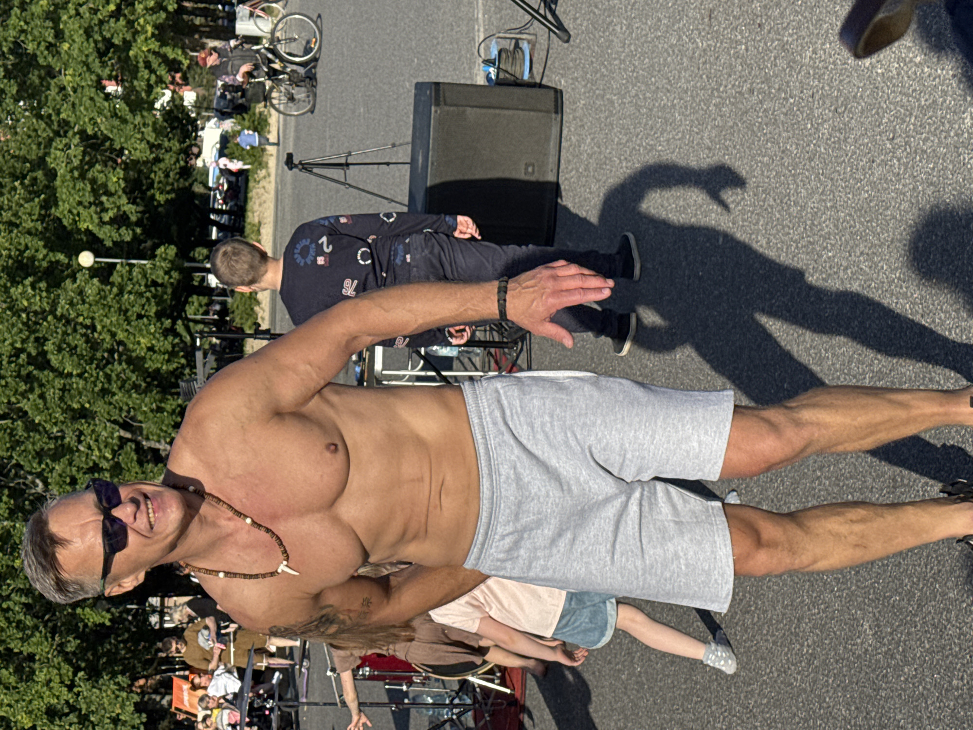 An outdoor, eye-level shot captures a shirtless man with tanned skin, wearing sunglasses and a bead necklace with a tooth pendant, as he stands in a city square. He is wearing light grey shorts and black sneakers. His muscular torso is well-defined, and his arms are raised slightly, with his right hand visible in the foreground. The man is smiling. Behind him, a young boy in a dark blue sweater and jeans stands facing away, near a large black speaker on a tripod. Further in the background, other people, bicycles, and trees are visible, suggesting an urban public space. The ground is paved with asphalt. The image has a shallow depth of field, with the man in the foreground in sharp focus. The lighting suggests a sunny day. Based on visual cues, this scene is consistent with Kadriorg Park in Tallinn, Estonia, possibly near the amphitheater or an open area where events are held.