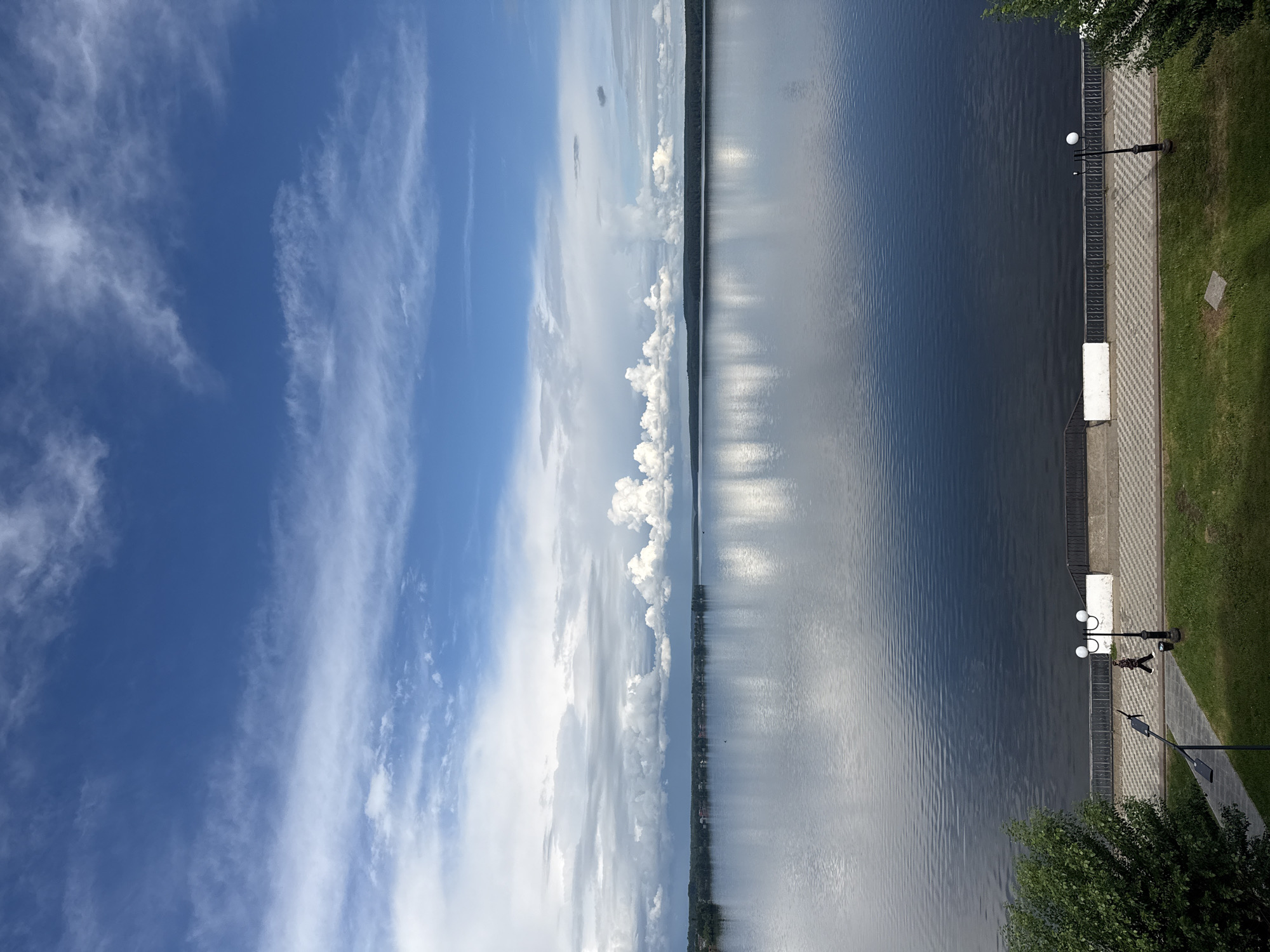 The image shows a view from an elevated position overlooking the Votkinsk Pond in Votkinsk, Russia. The left side of the image is dominated by a blue sky with scattered white clouds. A line of trees is visible in the middle distance, separating the sky from the water. The right side of the image depicts the dark, rippled surface of the pond. A paved walkway runs along the edge of the pond, bordered by grass and street lamps. A person is walking along the path. A bench is also present near the pond.
