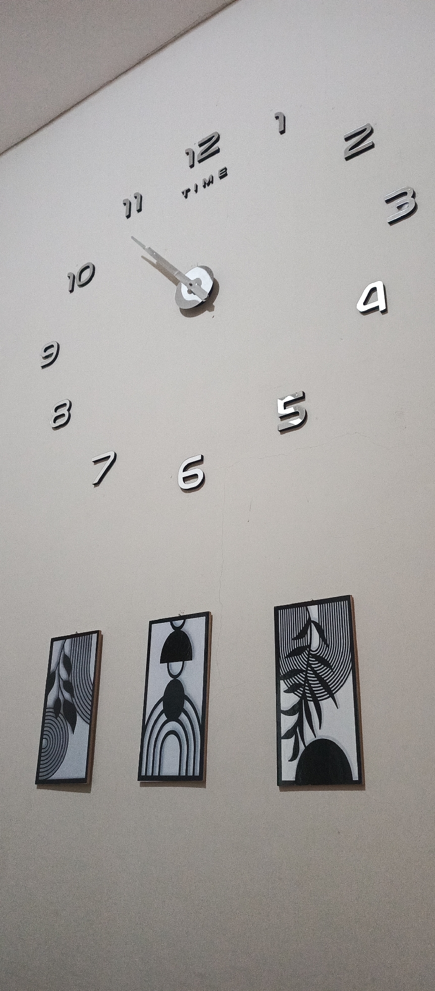 A wall-mounted clock with silver numbers is displayed on a beige wall. The clock has a silver hour hand pointing to the 10 and a silver minute hand pointing to the 11. Below the clock, three rectangular art pieces with black and white abstract designs are hung in a horizontal row. The art pieces appear to be in a home setting. The image does not provide any information about the location being Jatisari, Indonesia.