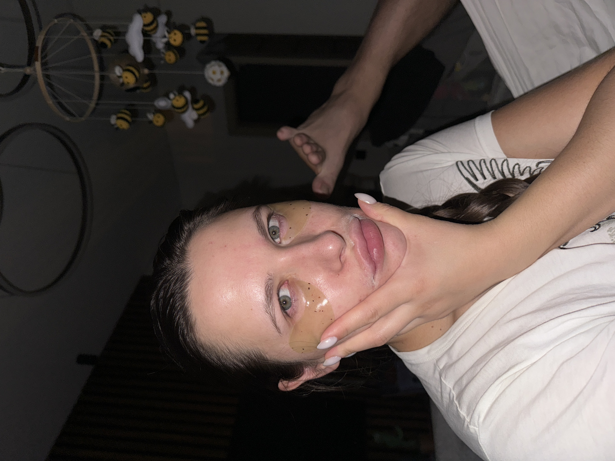 The image shows a person indoors with eye patches on, touching their face. A foot is visible in the background. In the background, a bee-themed mobile hangs from the ceiling. There is no information to indicate this picture was taken in Akulovo (Gp Kubinka), Russia.