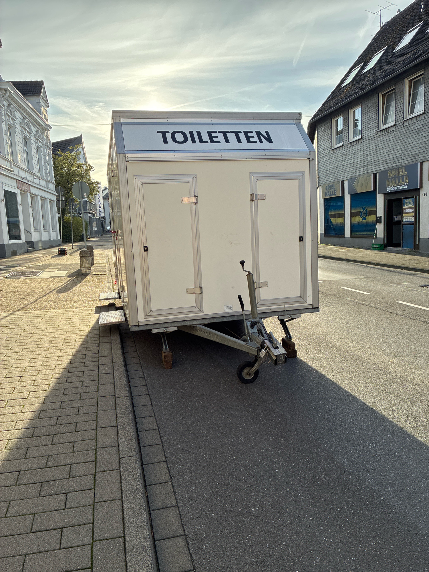 The image captures a street scene in Radevormwald, Germany, on a sunny day. The primary subject is a white portable toilet trailer positioned on the right side of a paved road, with its wheels on the asphalt. The trailer has two doors on its visible side and the word "TOILETTEN" is prominently displayed in black lettering on its top. Adjacent to the trailer, a sidewalk constructed of gray paving stones is visible. The street is lined with buildings on both sides. On the left, a white building with multiple windows and architectural details is present. On the right, a darker gray building features several windows and shops on the ground floor, one of which has signage reading "SPIEL HALLE RADEVORMWALD". The sun casts long shadows across the scene, indicating it is likely late afternoon or early morning. The sky is partly cloudy with visible contrails.