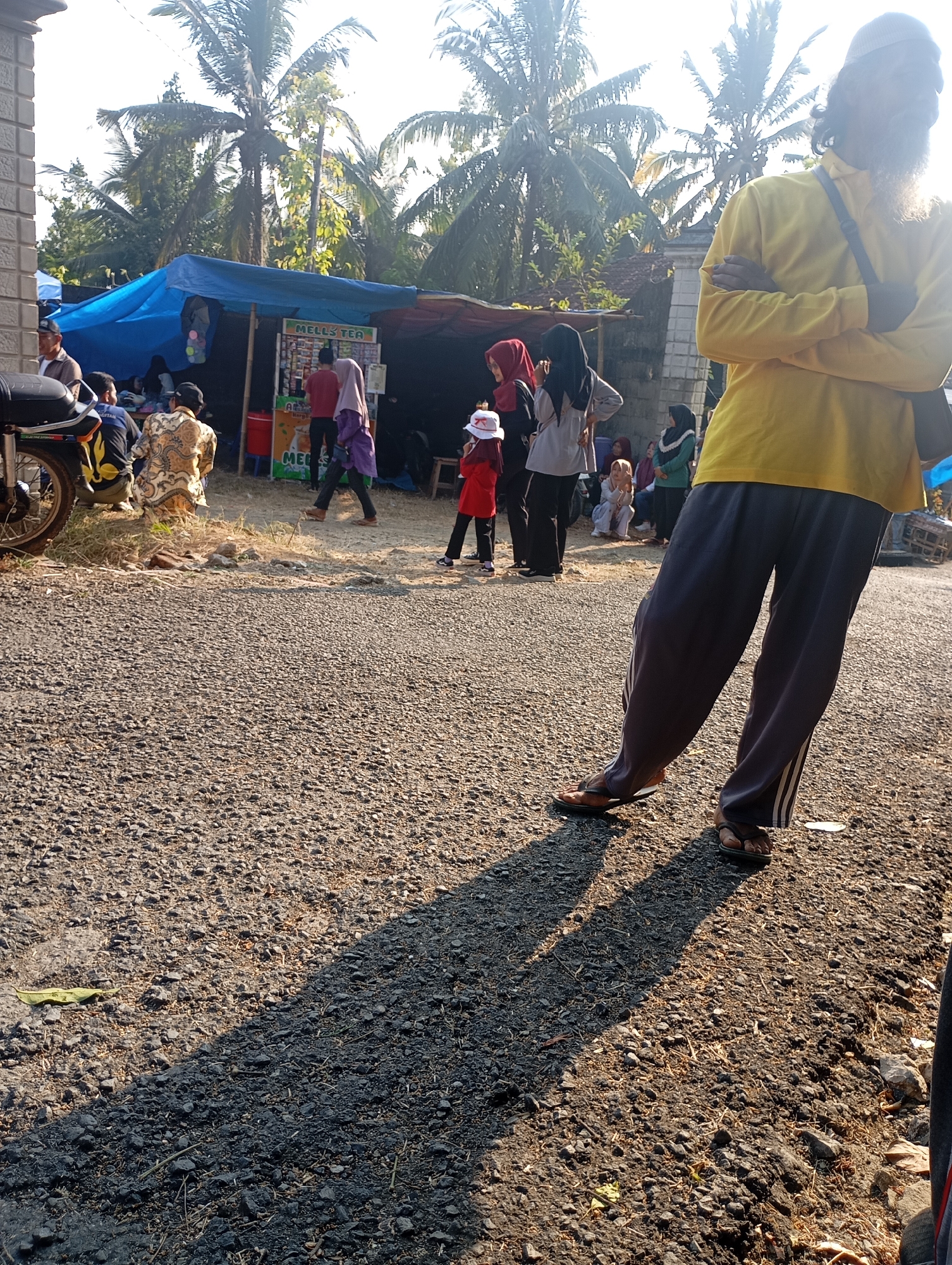 The image depicts an outdoor scene on a sunlit day, likely in Ketepung, Indonesia, with palm trees visible in the background. In the foreground, the legs of a person wearing dark pants and flip-flops are prominent, casting a long shadow on the gravelly ground. Behind this individual, several other people are gathered around a makeshift stall or tent with a blue tarp. The stall appears to be selling beverages, with signage that includes "MELL'S TEA". Some individuals are dressed in traditional Islamic attire, including headscarves. A motorcycle is parked to the left of the frame. The overall atmosphere suggests a casual gathering or local market.