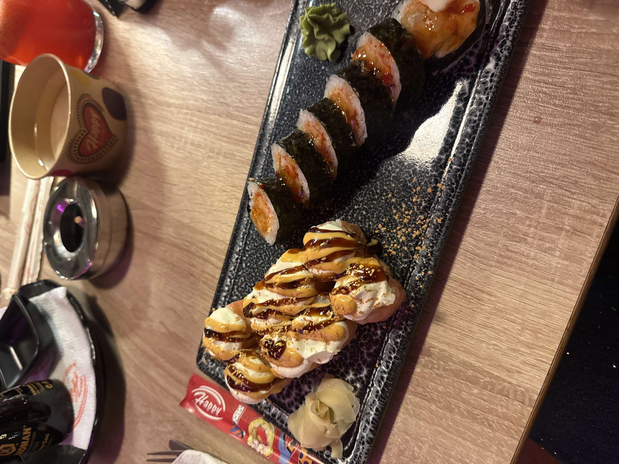 An overhead shot captures a dining table set with various dishes and accessories, likely in a restaurant or bar. The central focus is a long, dark speckled platter holding an assortment of sushi.
On the platter, a row of eight maki rolls, wrapped in dark nori seaweed, feature a light-colored filling, possibly shrimp or crab, topped with a glistening, reddish-brown sauce. Adjacent to these rolls is a dollop of green wasabi paste. Further down the platter, a cluster of irregularly shaped sushi pieces are topped with a generous amount of white cream, drizzled with a dark, glossy sauce, and sprinkled with what appear to be golden flakes. A small pile of pale yellow pickled ginger accompanies this section.
To the left of the platter, a clear glass filled with a bright red beverage, possibly juice or a cocktail, is visible. Below it, a beige ceramic cup features a large red heart design with the word "Happy" written inside and partially visible text "Adorés" or similar below the heart. A metallic ashtray with a dark object inside, resembling a cigarette butt, sits beneath the cup.
At the bottom left, a black bottle of "KIKKOMAN" soy sauce is seen, along with a pair of chopsticks resting on a red wrapper that also says "Happy". A fork is partially visible to the right of the chopsticks. The table surface is made of light-colored wood or laminate. The overall lighting suggests an indoor, evening setting.