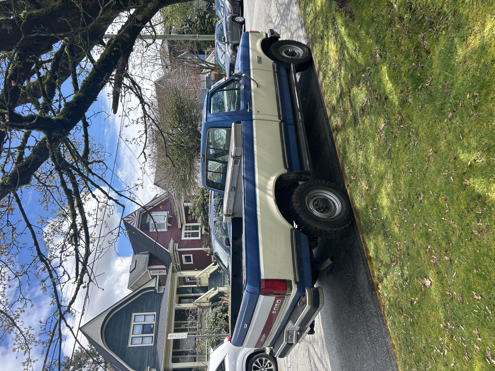 The image shows a blue and white Ford pickup truck parked on a residential street. The truck appears to be an older model with a toolbox in the bed. The houses in the background are Victorian-style, a common architectural style in Vancouver, Canada. A tree with moss-covered branches overhangs the street, casting shadows on the lawn beside the road. The sky is blue with scattered clouds. Other cars are parked further down the street. The scene suggests a typical residential area in Vancouver.