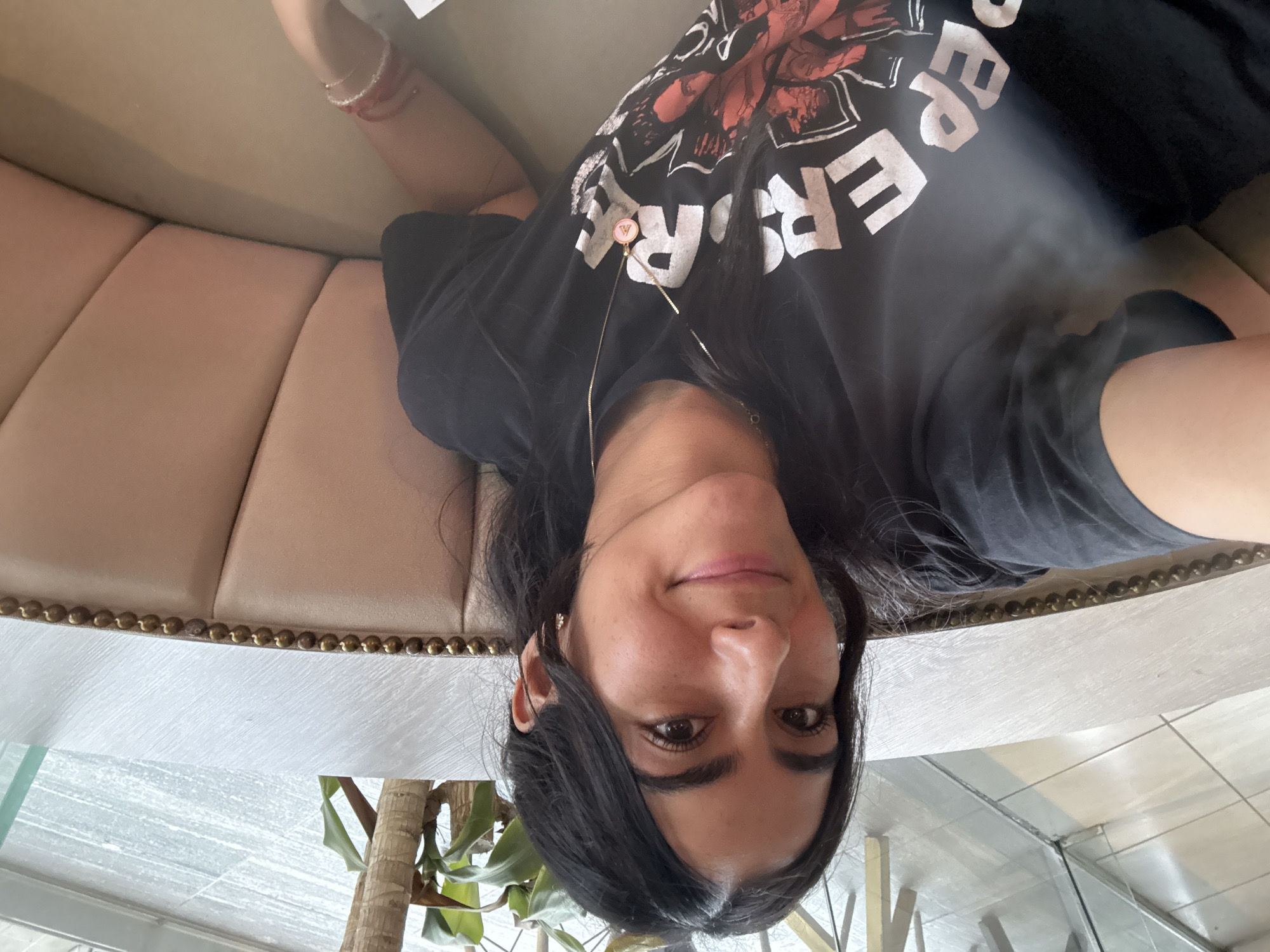 A woman is sitting inside a building, possibly a restaurant, in Puebla, Mexico. She is wearing a black t-shirt with white text and a red and white graphic, as well as a thin gold necklace. She is sitting on a booth with tan upholstery and decorative brass studs along the edge. A potted plant is visible in the lower left corner of the frame. The floor is tiled in a light-colored pattern. A glass partition is also visible.