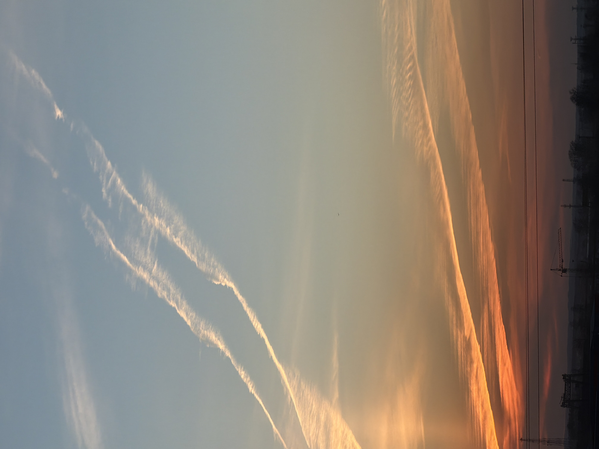 The image depicts a sunset scene over Chaikovsky, Russia, evidenced by the faint urban skyline and industrial cranes silhouetted against the horizon. The sky exhibits a gradient of colors, transitioning from a pale blue in the upper left to a warm orange near the horizon. Prominent contrails, likely from aircraft, stretch across the sky. Two distinct sets of contrails are visible in the blue portion of the sky, appearing as white or light-colored streaks. Near the horizon, the contrails take on a reddish-orange hue, mirroring the colors of the setting sun. In the lower right, a thin layer of trees and power lines can be seen along the horizon. The overall impression is that of a partially industrialized area under a sky marked by both natural beauty and the signs of air travel.