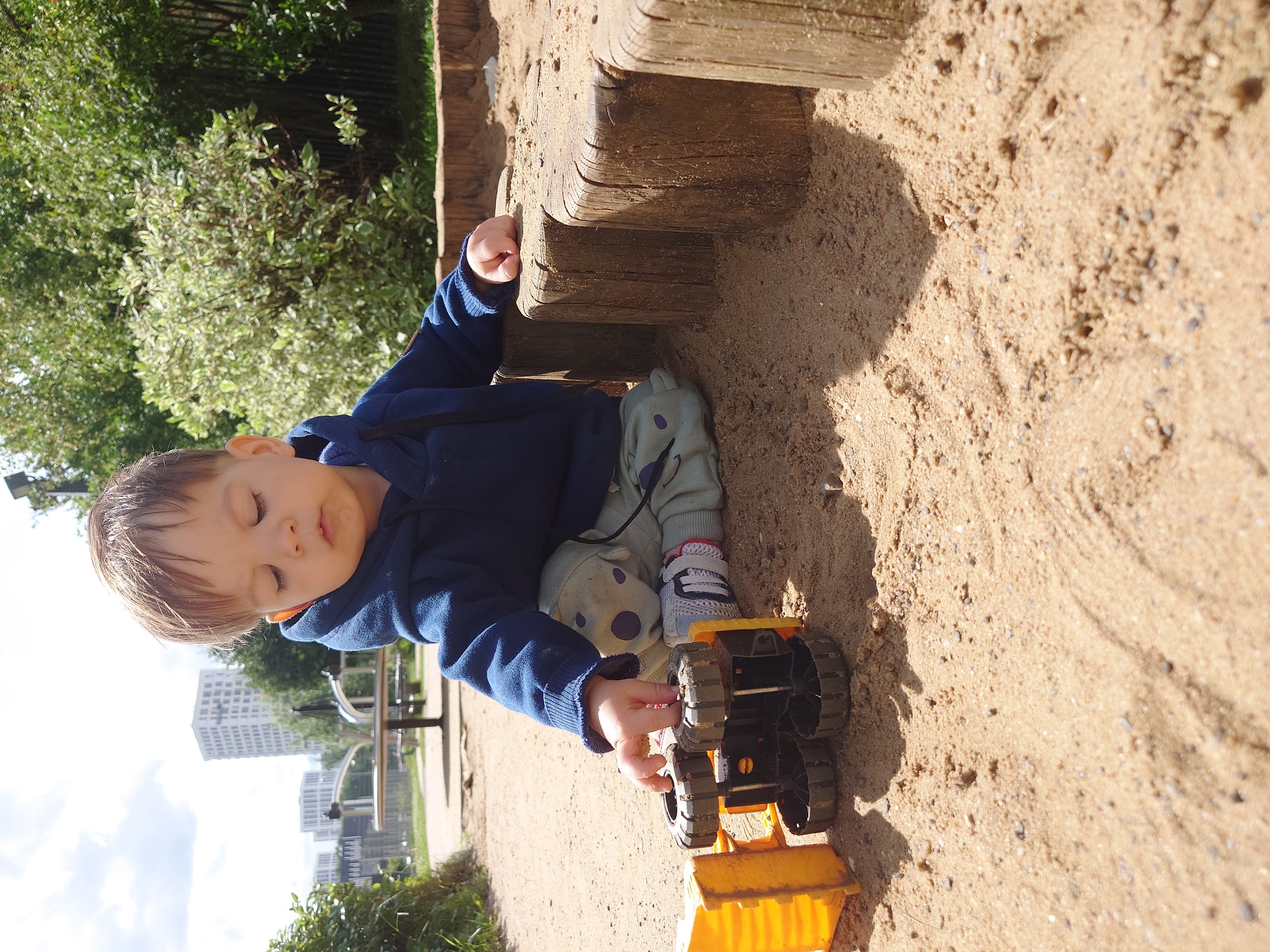 A young child, likely a toddler, is seated in a sandbox playing with a yellow toy truck. The child is wearing a navy blue hooded sweatshirt and light green pants with a pattern of purple dots. The sandbox is bordered by a series of wooden posts. In the background, there is a playground structure, lush green trees, and modern apartment buildings. The scene appears to be in a park or playground in the city of Kommunarka, Russia, given the context of the location. The lighting suggests it is daytime with natural sunlight casting shadows.