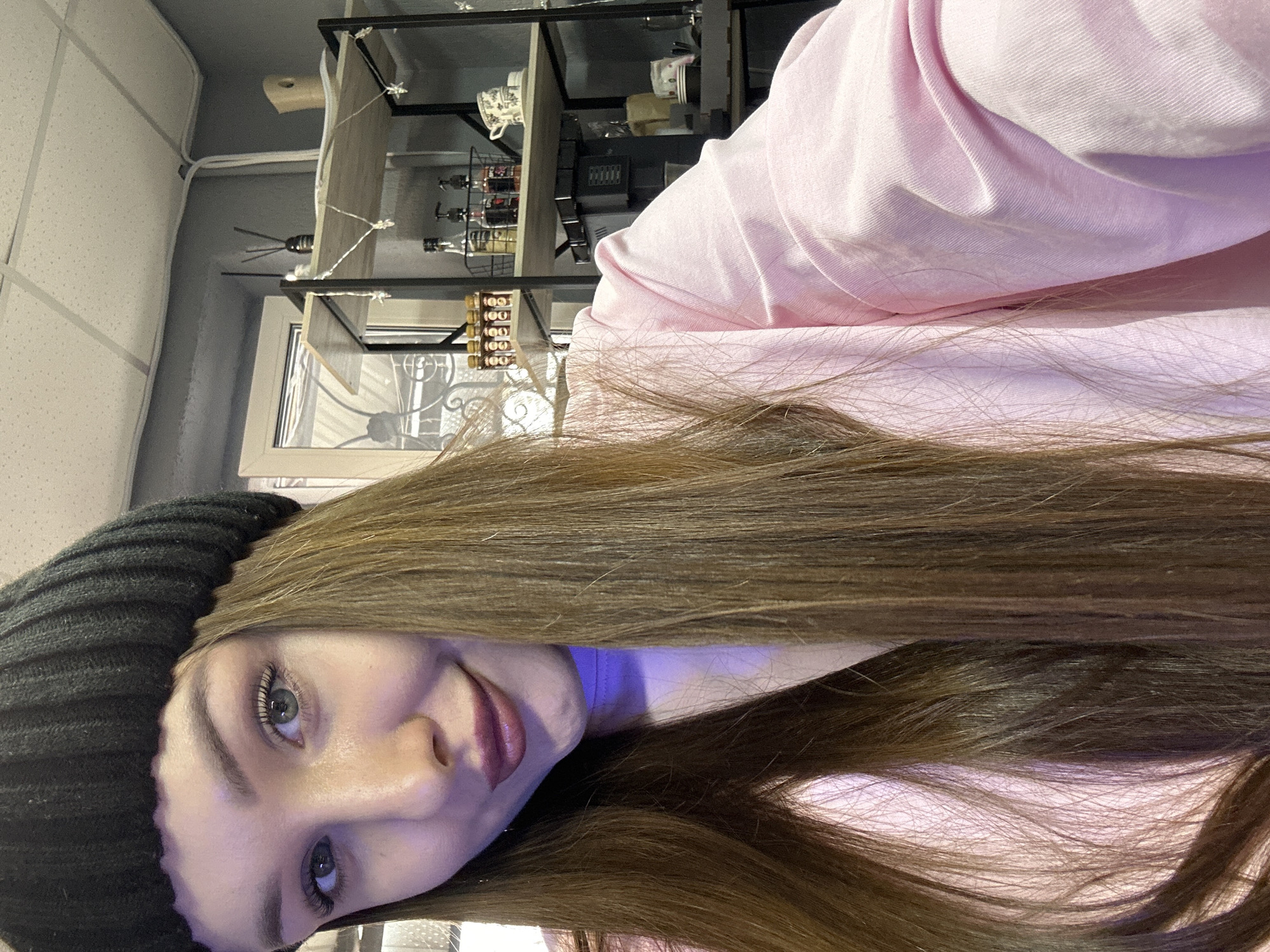 A young female is depicted in a close-up, front-facing indoor shot, positioned to the left of the frame. She wears a black knitted beanie, a light pink long-sleeved shirt over a purple undergarment, and has long brown hair. Her eyes are light-colored, and she exhibits a slight smile with dark pink lipstick. The setting is an interior commercial space in Chaikovsky, Russia, characterized by a gray wall and a white grid-panel ceiling. A white-framed window with exterior metalwork is visible on the left. To the right, a black-framed shelving unit with light wood-toned shelves holds various items: a black reed diffuser and a light-colored object adorned with star-shaped string lights on the top shelf; a metal basket with three pump bottles, a stack of two patterned cups, and a line of small amber bottles on the middle shelf. Below, part of a beverage machine and disposable cups are present. White electrical conduit is visible along the upper wall.