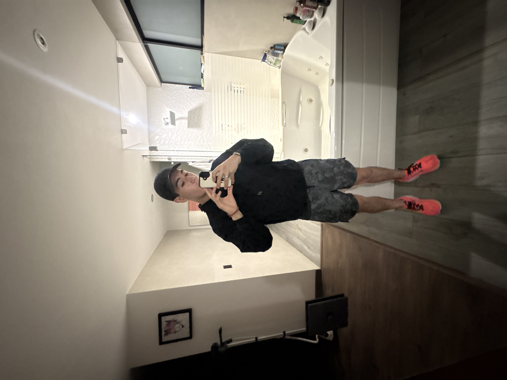 This media file captures a mirror selfie taken by a young person in a modern interior setting, located in San Bernardino Tlaxcalancingo, Mexico. The image is rotated 90 degrees counter-clockwise from its natural upright orientation.
The subject is a young person, possibly male, wearing a black baseball cap, a black long-sleeved shirt, and dark patterned shorts. They are holding a smartphone with a light-colored case up to their face to take the photograph. Their bright orange/pink athletic shoes with black detailing are prominent.
The background of the selfie (the area behind the person in the mirror) reveals a contemporary bathroom. This includes a white jacuzzi or large bathtub on the right, with several bottles of toiletries placed on its rim. A shower enclosure with clear glass panels is visible, along with a white textured tiled wall featuring a wavy pattern. A metallic ventilation grate is also present on this wall. Recessed ceiling lights illuminate the bathroom.
In the foreground of the image (the actual space where the person is standing), which is reflected in the mirror, there is a low white platform or bed. A small, framed artwork hangs on the wall next to it. Further to the right, a floor stand with a black monitor or screen is visible. The flooring throughout the visible areas consists of dark wood-look planks or tiles.
The activity depicted is the casual act of taking a mirror selfie. The setting appears to be a private room or suite, characterized by clean lines and modern finishes. The lighting is artificial and bright. No discernible text is visible on any objects or clothing.
