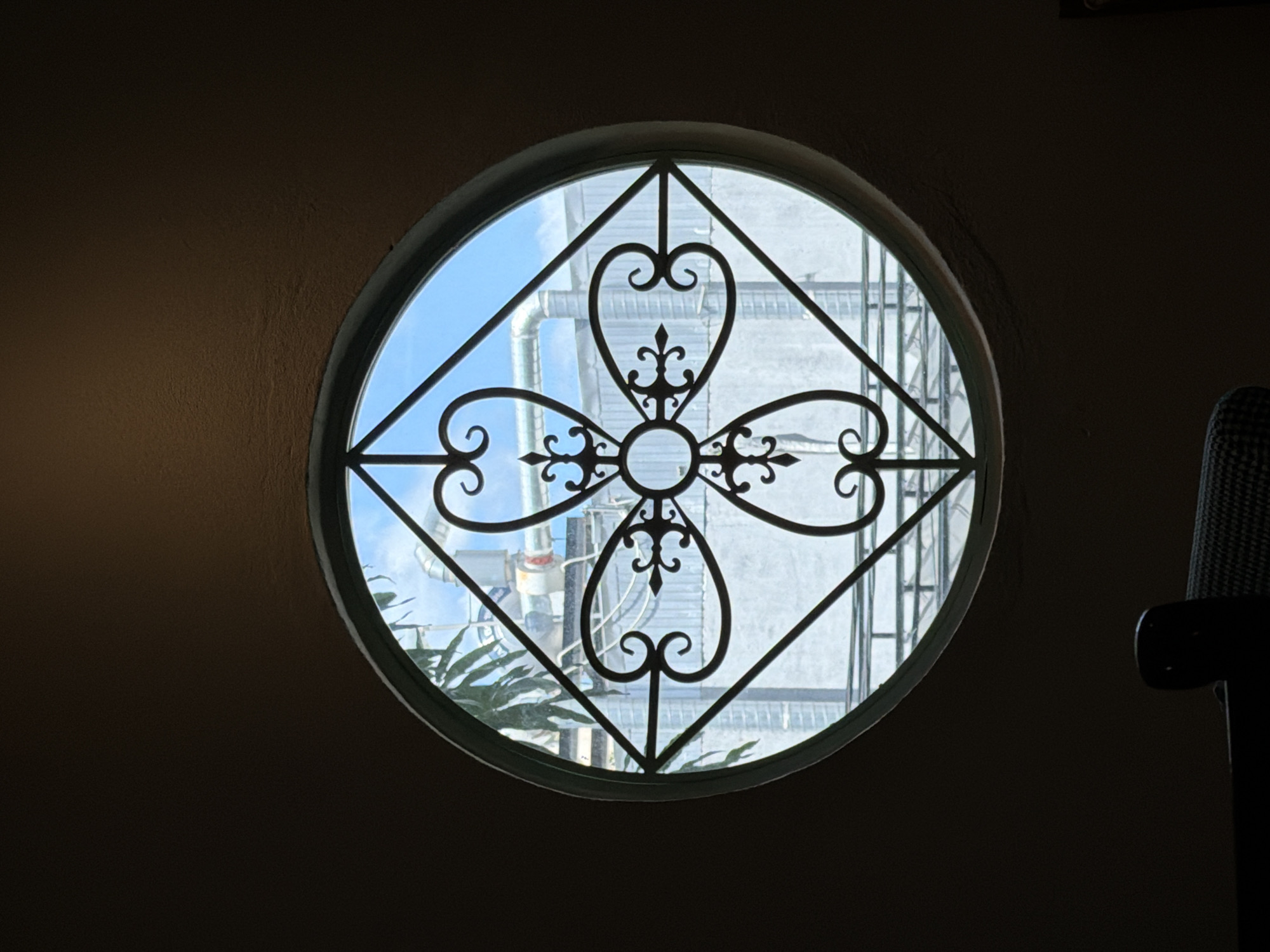 The image depicts a circular window with an ornate wrought iron grill in the foreground, set against a dark wall. Through the window, a bright blue sky is visible, partially obscured by an industrial structure with pipes and ventilation systems. A palm frond from a nearby tree can be seen in the upper left quadrant of the window. The scene is likely captured from an interior space looking outwards, possibly in the city of Đà Nẵng, Vietnam, given the architectural style and the presence of industrial elements often found in developing urban areas.
