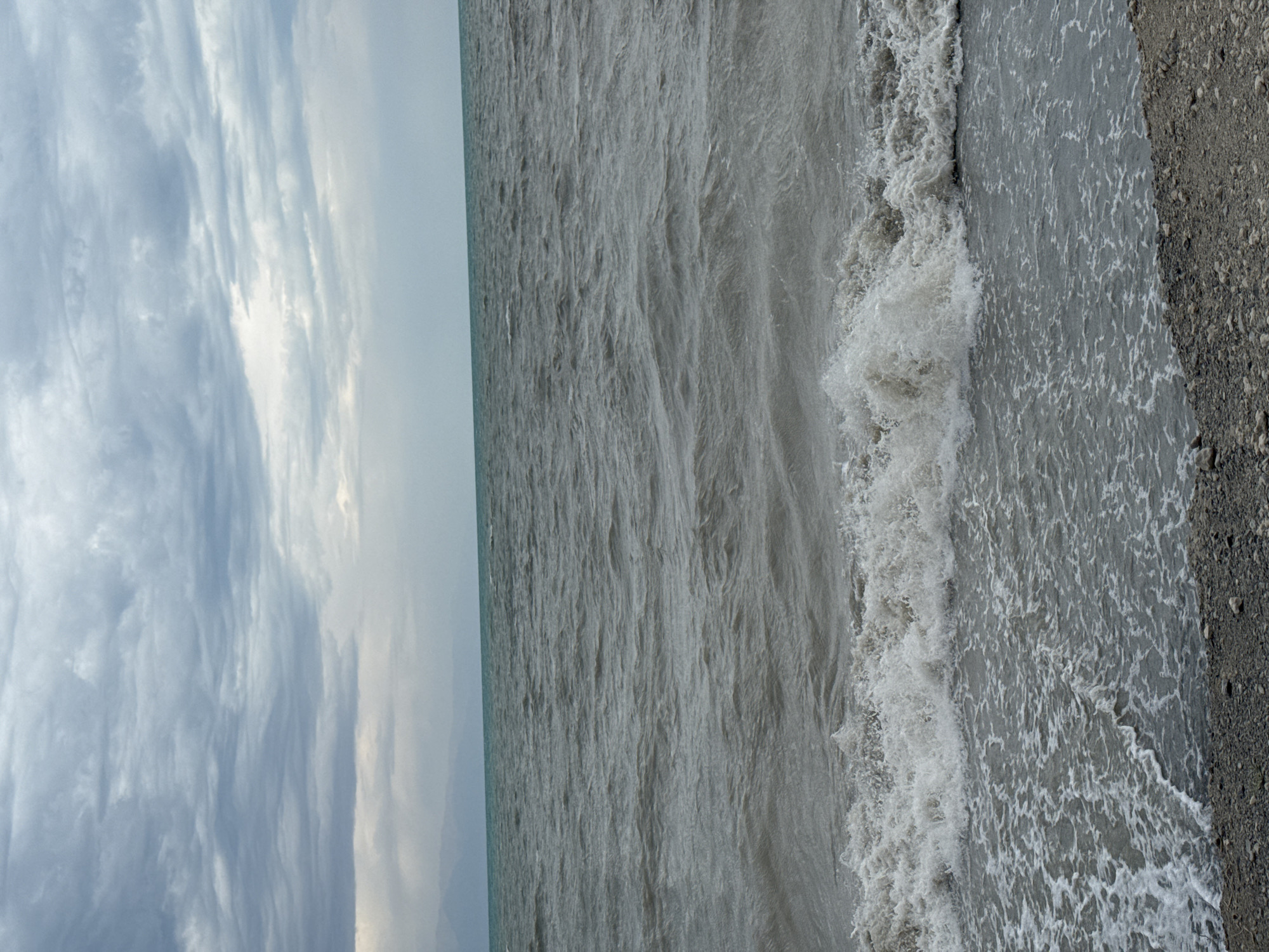 The image depicts a cloudy day at a body of water, likely a lake or sea, with waves crashing onto a rocky shore. The water is a murky grey-green, and the sky is filled with heavy, layered clouds. In the distance, faint mountains are visible on the horizon. The foreground shows a pebbled beach with small rocks and some scattered debris. The overall impression is one of a dramatic, overcast natural scene. This location is identified as Urdzhar, Kazakhstan.