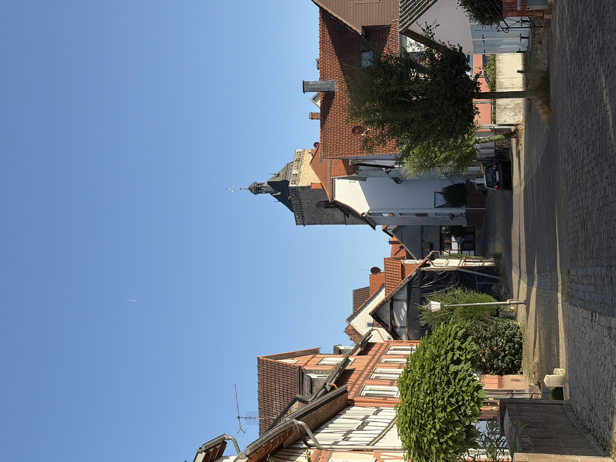 The image depicts a narrow, cobblestone street in a European town. On the left, traditional half-timbered houses with red-tiled roofs line the street. A lush green tree with a rounded canopy is prominently featured in the foreground, casting a shadow on the street. A stone wall separates the sidewalk from the street on the left. A lamppost stands on the sidewalk. Further down the street, a church tower with a dark roof and cross dominates the skyline. The sky is clear blue with a faint contrail visible. On the right side of the street, more houses with red-tiled roofs are visible, some with white facades. A dark-colored car is parked on the right side of the street. The overall impression is of a quiet, historic town during daylight hours.