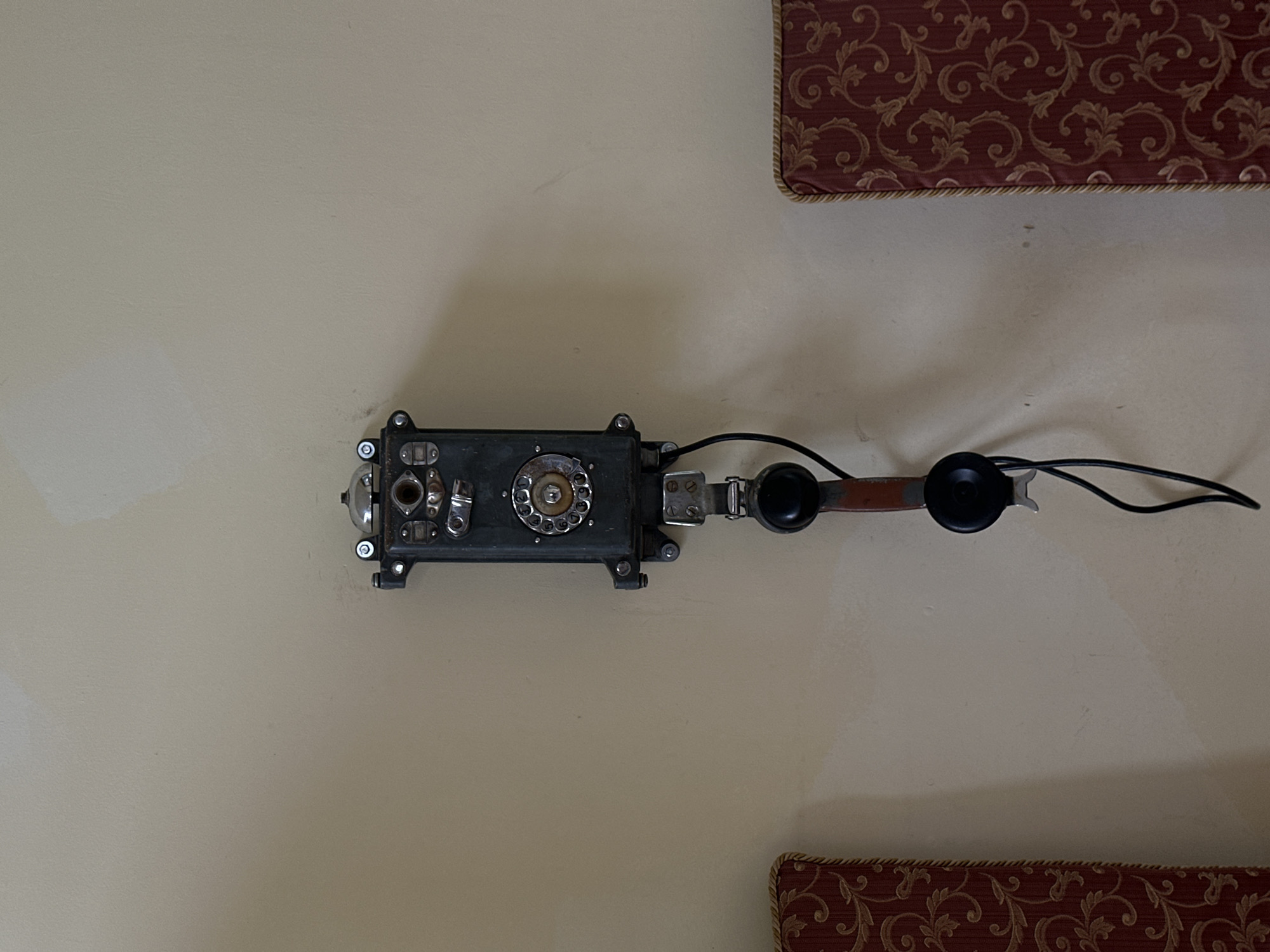 A vintage, wall-mounted telephone, predominantly black with some metallic components, is affixed to a light beige wall. The telephone features a rotary dial and a separate earpiece/microphone connected by a cord. Red and gold patterned upholstery is partially visible in the upper right and lower right corners of the frame. There is no indication of the location of this image being in Sochi, Russia, but it appears to be in some building.