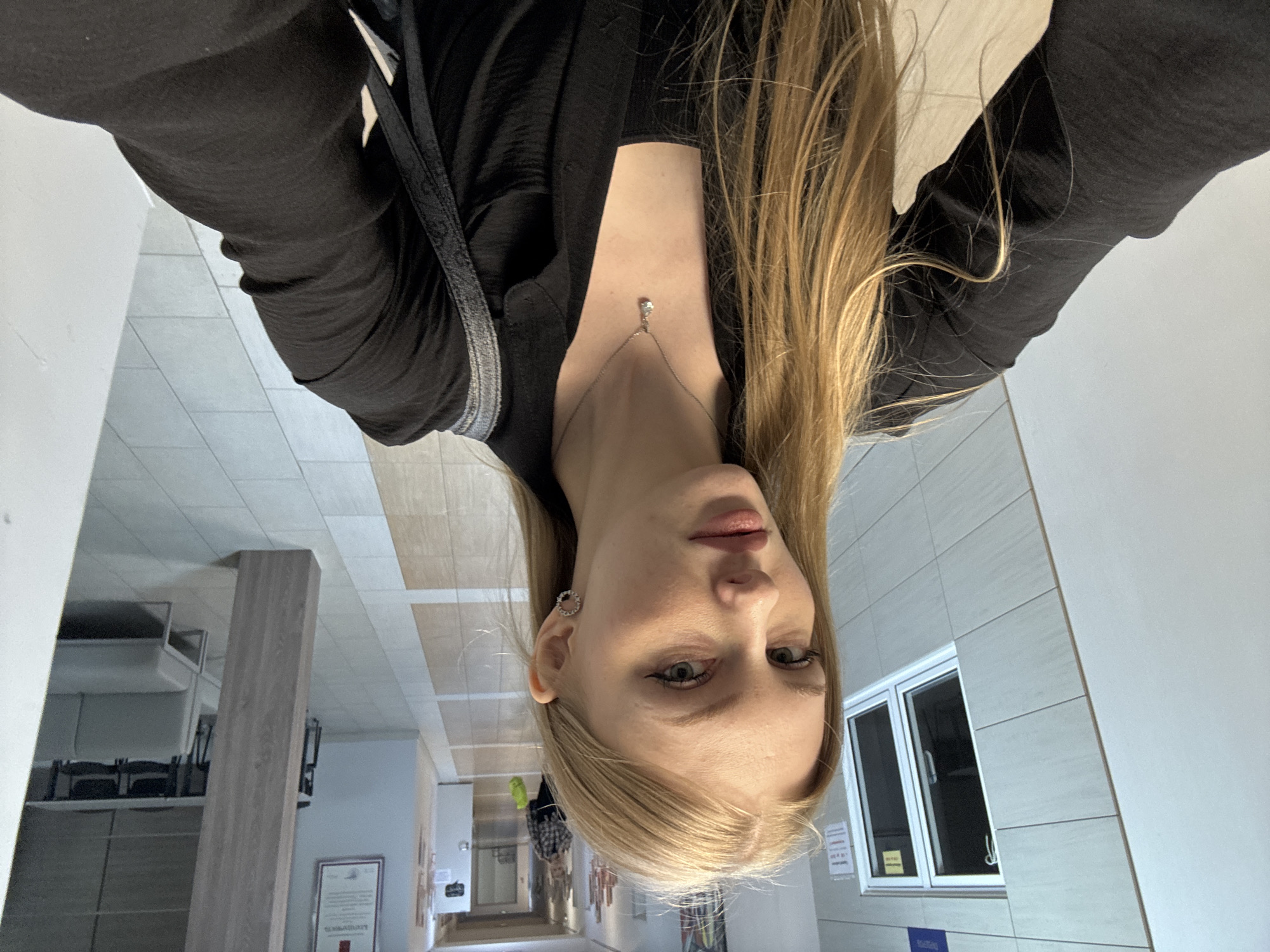 A woman with blonde hair is taking a selfie indoors. She is wearing a black top and a necklace with a pendant. The background shows a brightly lit hallway with tiled floors and a wall with windows. A display case and informational posters are visible along the hallway. The location appears to be an institutional or office building. The available image data does not contain information confirming the location as Perm, Russia.