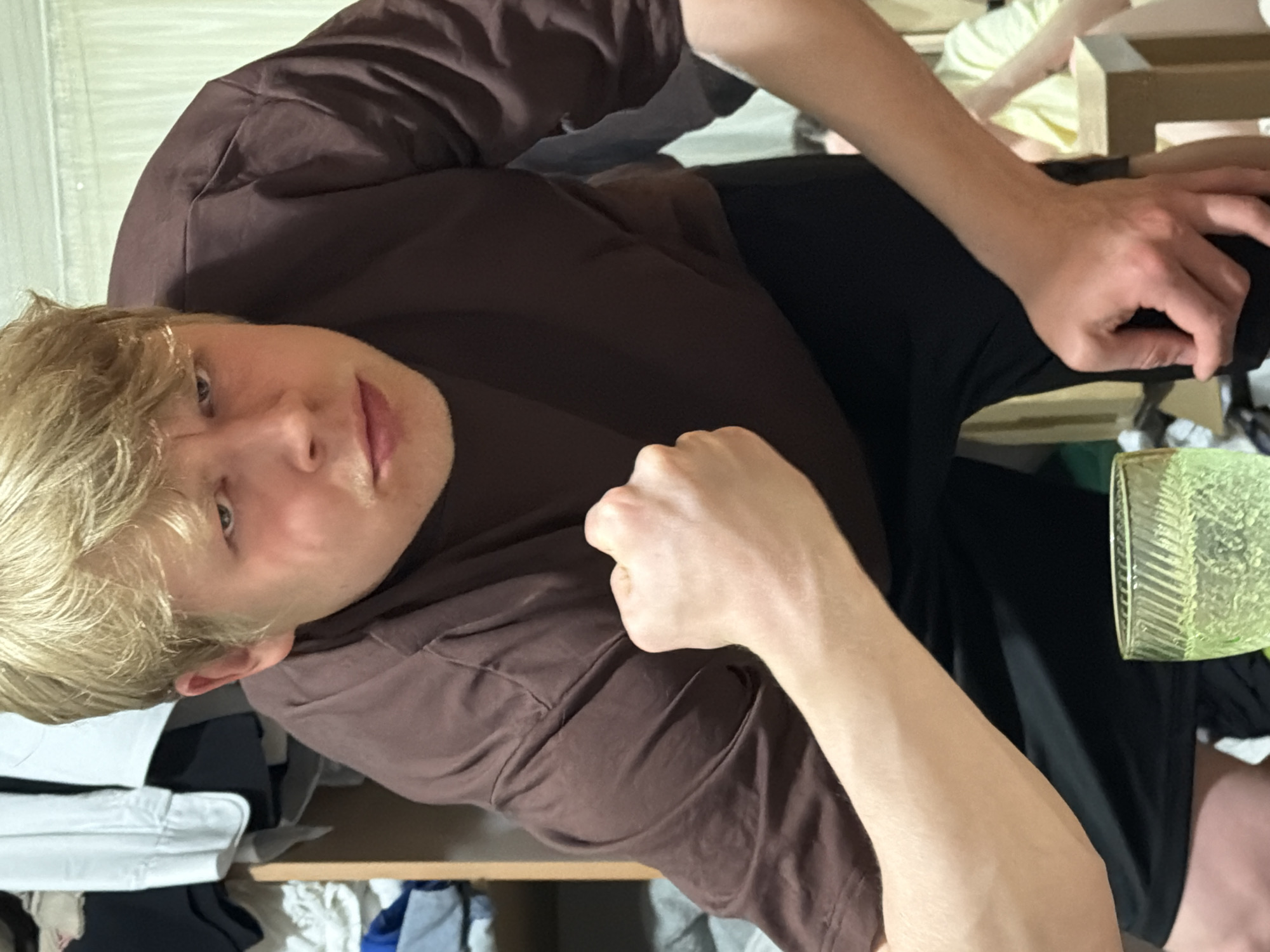 A blonde-haired person wearing a brown t-shirt and black shorts is positioned in what appears to be an indoor setting, possibly a bedroom or closet. The camera angle is looking up towards the person. A transparent textured glass is visible near the lower right corner. Cluttered clothing and storage units are observed in the background. The specific location within Izhevsk, Russia, cannot be determined from the image.
