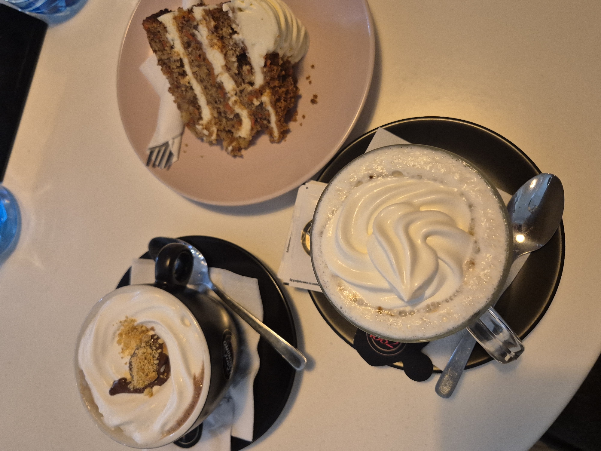 The image shows a tabletop scene in a cafe. A slice of carrot cake with white frosting sits on a pink plate in the upper left quadrant. Two beverages in glass mugs are positioned below and to the right of the cake. The mug on the left is black with a handle and topped with white whipped cream, chocolate sauce, and crumbly toppings. It is placed on a black saucer. The second mug, closer to the center, is transparent with a handle and contains a frothy beverage topped with a swirl of white whipped cream, set on a black saucer. A silver spoon rests across the top of the saucer and mug. Napkins are placed beneath the cups. The background is a plain, light-colored tabletop. A blue water bottle is partially visible in the top left corner. The location is Pavlikeni, Bulgaria, as indicated by the cafe's logo on a small black coaster.