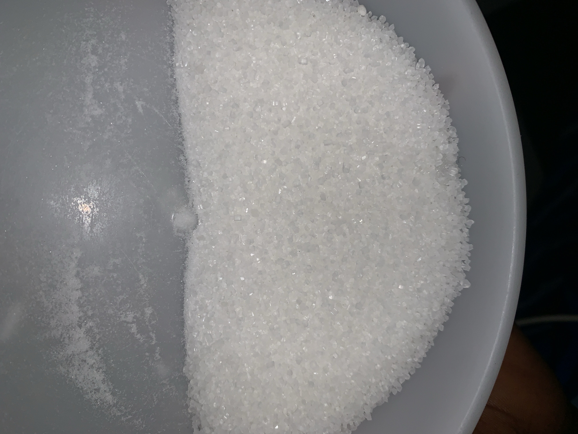 A portion of a white, possibly plastic, bowl is filled with white granular material. One half of the bowl appears empty, while the other is densely packed with the grains. The granular material resembles sugar. The setting and location in Rafin Par, Nigeria, are not ascertainable from the image.