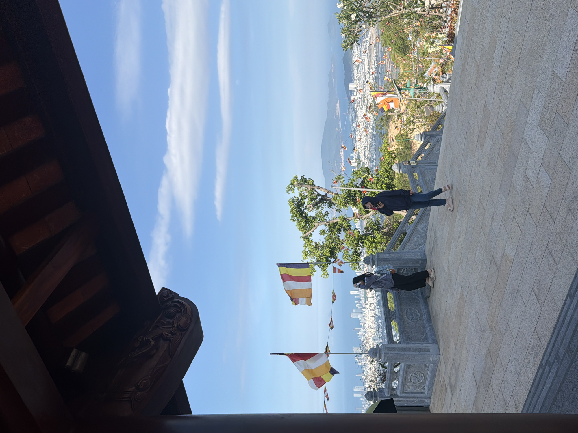 The image shows a view from a high vantage point, possibly a temple or elevated structure, overlooking the city of Nam Nha Trang Ward, Vietnam. Two people stand on a stone-paved area, one appearing to be using a phone, both looking out at the city. Buddhist flags are visible, indicating the location's religious significance. The sky is blue with light clouds, and the cityscape extends to the coast. A portion of a dark, ornate roof structure is visible in the foreground. The overall environment suggests a tourist destination with a scenic overlook.