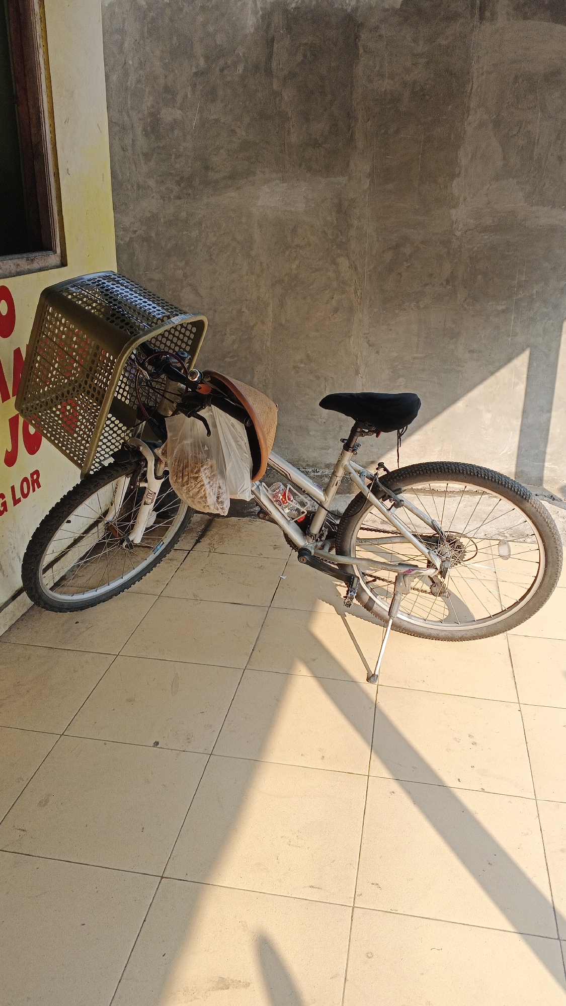 An outdoor, eye-level shot captures a silver bicycle with a large, woven basket attached to the handlebars. The basket is partially filled with a clear plastic bag containing what appears to be dried goods. A brown wicker-like piece of material is affixed to the front of the handlebars. The bicycle is parked on a tiled floor, and a kickstand supports it. Behind the bicycle is a textured, concrete wall that casts shadows from an unseen light source. To the left of the bicycle, a portion of a yellow wall with red lettering is visible. The scene is illuminated by bright sunlight, creating distinct shadows on the tiled ground. The location is identified as Ambarketawang, Indonesia.