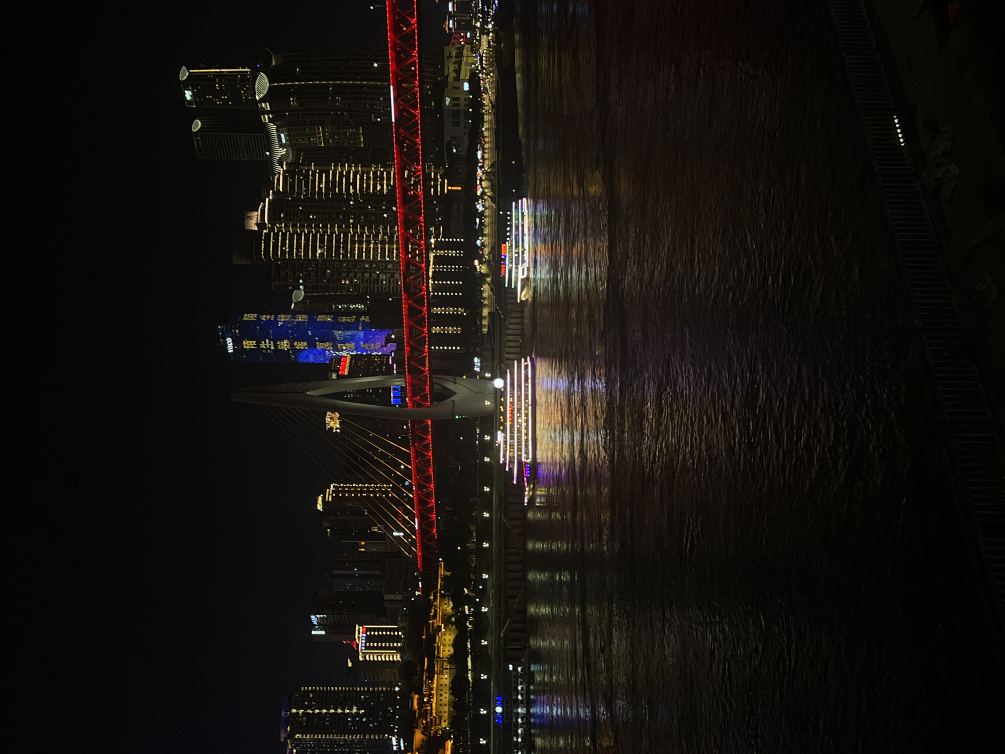 This is a nighttime cityscape photograph taken in Nan'an District, Chongqing, China. The image features a wide river in the foreground, reflecting the colorful lights of the city and two illuminated boats. In the middle ground, a distinctive cable-stayed bridge spans the river, adorned with vibrant red lighting. Behind the bridge, a cluster of modern skyscrapers dominates the skyline, their windows lit with warm golden light. One tall building on the left displays blue lighting with Chinese characters and the "YOCO" logo. The dark night sky is visible above the city lights. The overall scene depicts a bustling urban environment at night, characterized by impressive architecture and illuminated waterways.