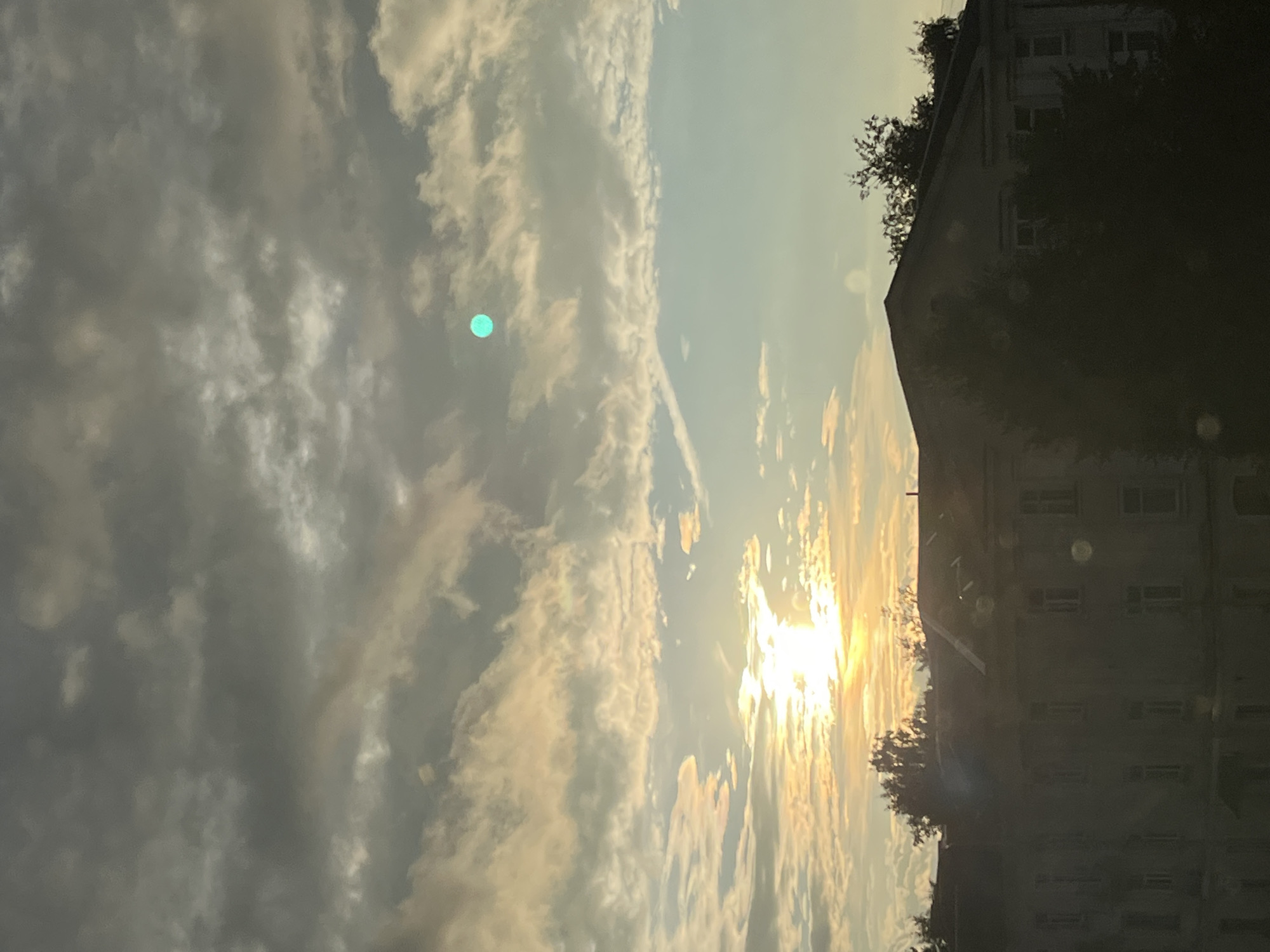 A photograph taken from a low angle shows the sky above a building and trees. The sky is filled with clouds, some dark and heavy, others lighter and wispy. The sun is setting or rising, casting a warm glow on the clouds and creating a bright light in the center of the image. A small, bright teal disc is visible in the upper right portion of the sky. The building in the foreground appears to be old and somewhat dilapidated, with visible windows and architectural details. Trees with dark foliage obscure parts of the building. The overall scene suggests a late afternoon or early morning light over an urban landscape. The location is identified as Kohtla-Järve linn, Estonia. There are no people visible in the image.