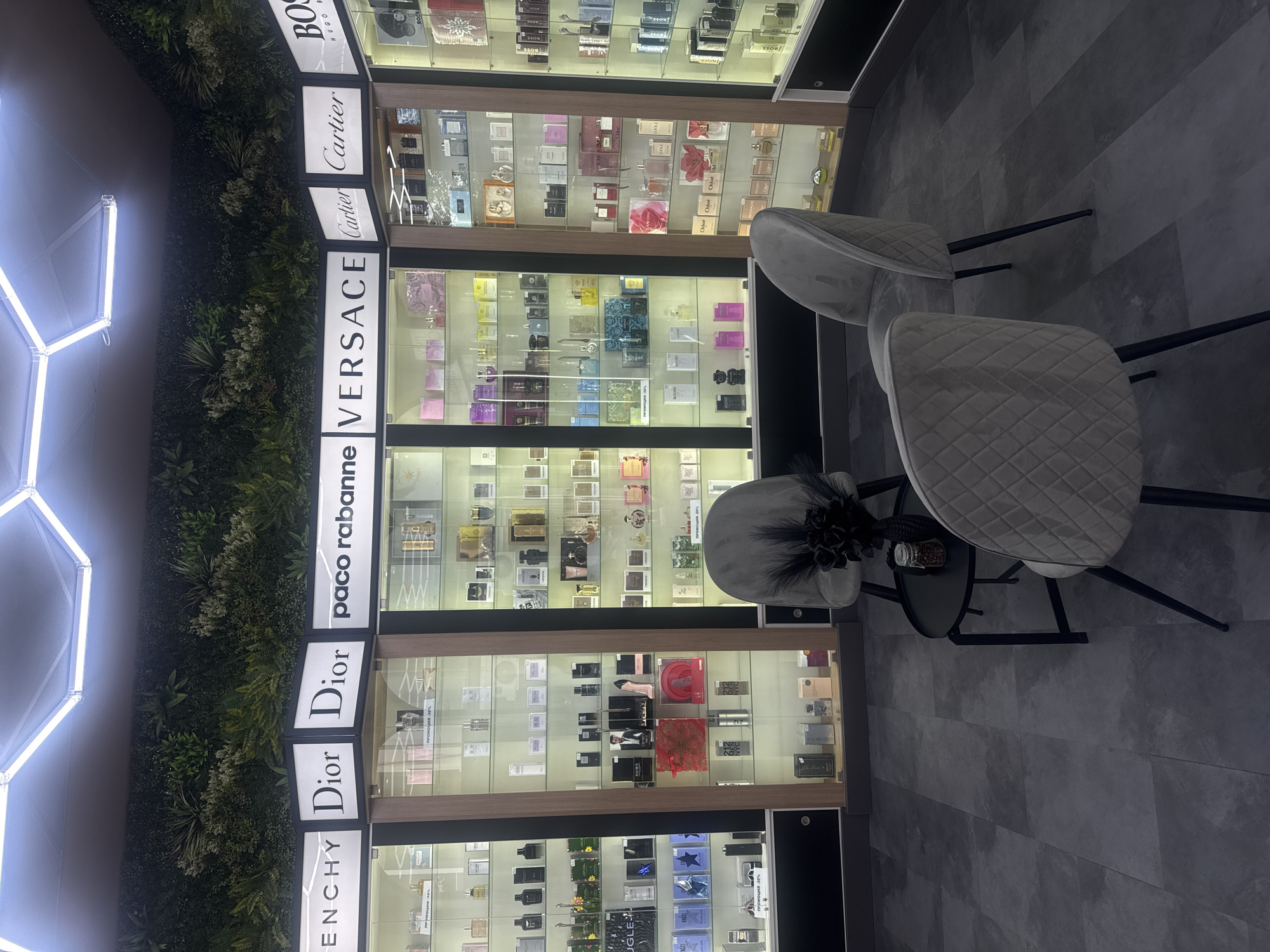 The image shows the interior of a retail space, likely a perfumery. Cabinets displaying various perfume brands (Givenchy, Dior, Paco Rabanne, Versace, Cartier, Hugo Boss) are visible. Two chairs and a small table with decorative objects are also present. The lighting is bright, and the floor is tiled in a grey tone. The location of the image could be Kazanlak, Bulgaria, although this cannot be definitively confirmed from the image alone.