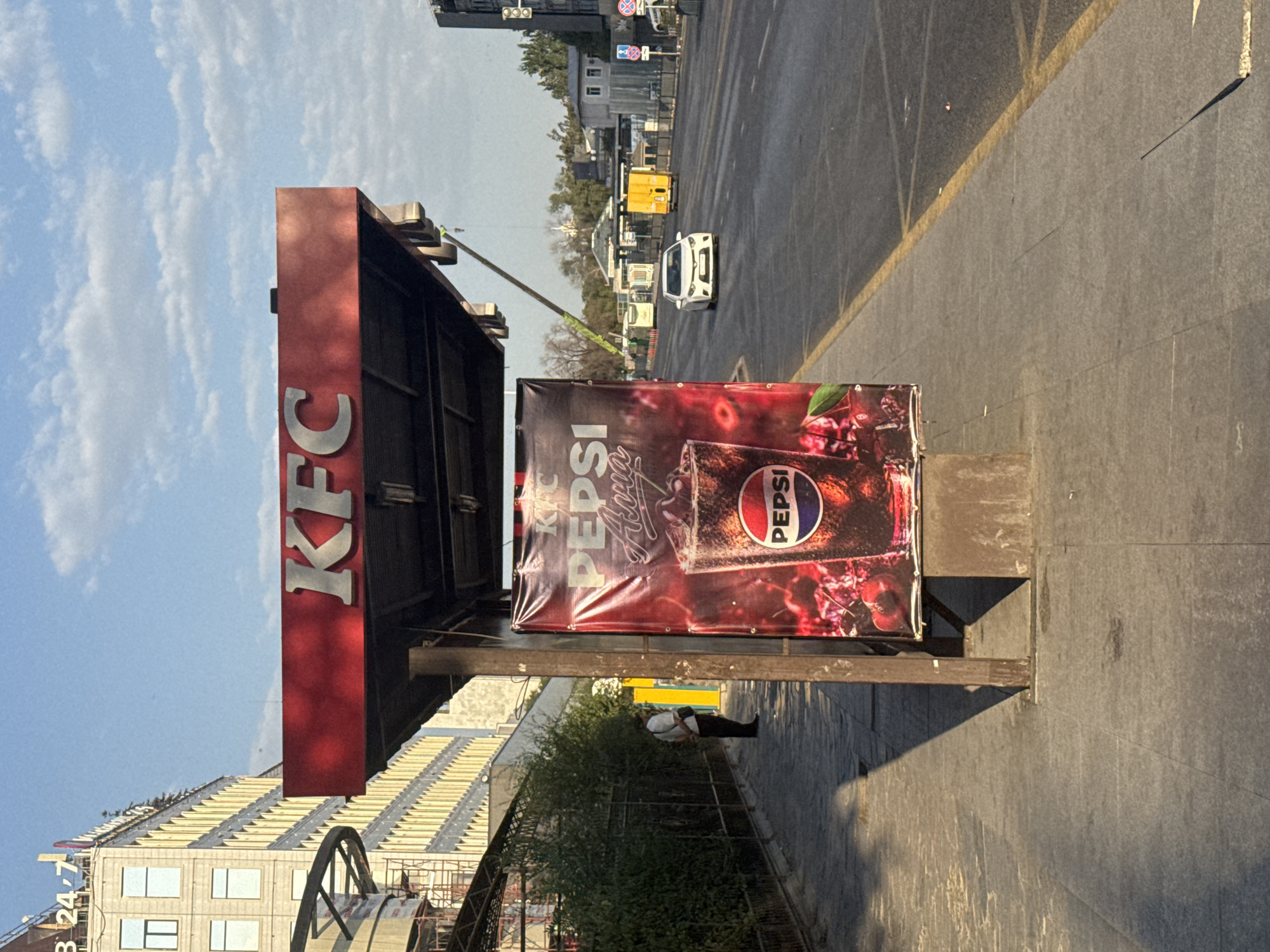 A Pepsi advertisement is displayed on the side of a KFC bus stop shelter in Bishkek, Kyrgyzstan. The ad features a glass of Pepsi with cherries. A person is standing at the bus stop, facing away from the camera. The bus stop is located on a city street with traffic and buildings in the background. The sky is blue with some clouds.