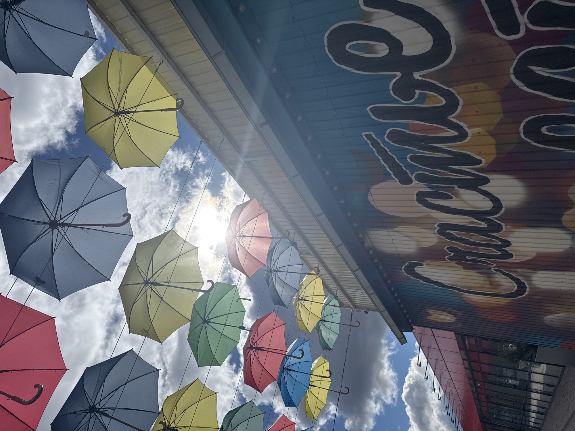The image shows a view looking up at a display of colorful open umbrellas hanging in the air. Colors include yellow, blue, red, green, and grey. The umbrellas are arranged in a pattern against a blue sky with white clouds. The sun shines through the umbrellas. To the right of the umbrellas is a building with artwork or signage on the facade. The signage features the word "Chacha" in cursive. The location is indicated to be Osa, Russia. There are no people visible.