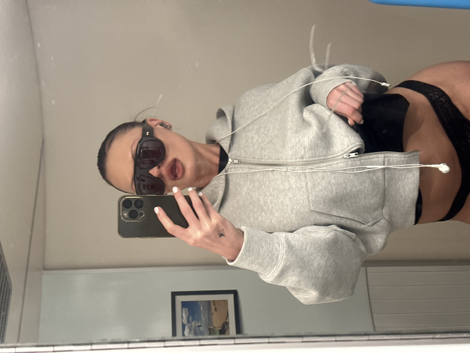 The image depicts an adult female taking a mirror selfie in an indoor setting. The specific location in London, United Kingdom, cannot be definitively determined from the visual content. The subject, facing the mirror, holds a dark smartphone with a prominent multi-lens camera array in her right hand to capture the reflection. She wears oversized, dark, wraparound sunglasses, and her lips are darkly colored. Her hair is pulled back from her face. She is dressed in a light grey, cropped, full-zip hoodie, unzipped to reveal a black crop top or bra and a black lace thong. Wired white earphones hang from her ears and drape across her torso. Her fingernails are painted white, and a small dark tattoo, resembling a star, is visible on the back of her left hand. The background reflection shows plain, light-colored walls. On the left, a framed picture depicts a beach scene with a person on a sandy shore under a cloudy sky. Vertical white tiles are partially visible on the left edge, and a ceiling light fixture is discernible at the top.