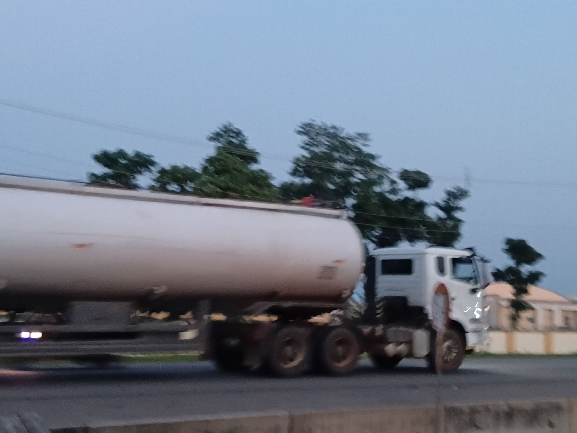 The image shows a white fuel tanker truck driving on a road in Jalingo, Nigeria. The truck is in motion, as indicated by the motion blur. The truck has a large cylindrical tank and a white cab. The background includes trees and buildings, suggesting an urban or semi-urban environment. The sky is overcast and the lighting suggests it is either early morning or late evening. The road appears to be paved, and there is a concrete barrier on the side of the road. The overall scene depicts typical road traffic in Jalingo.