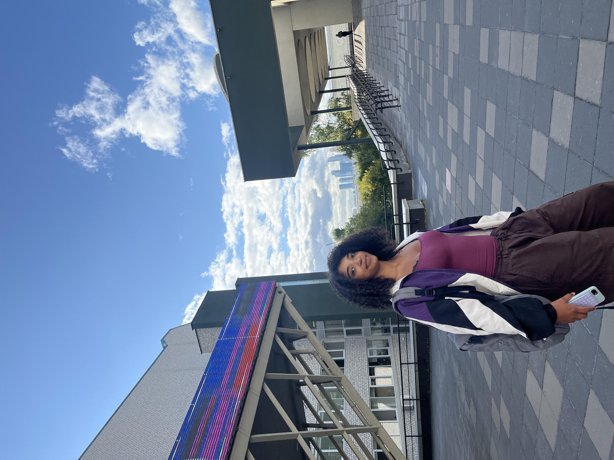 A young Black woman with curly hair stands on a patterned tiled walkway under a blue sky with white clouds. She is wearing a purple tank top, a multi-colored jacket with purple, white, and black sections, and brown pants. A grey backpack is on her shoulders, and she holds a light purple phone in her right hand. Behind her, a modern building with a large, colorful LED display is visible, along with an overpass structure. In the distance, a cityscape with several skyscrapers can be seen, with trees and foliage in the foreground. The architectural style of the buildings and the distant skyscrapers suggest the location is likely in a modern urban area of Moscow, Russia, possibly near a university or cultural complex. A person is faintly visible in the distance on the right, walking along a covered walkway.