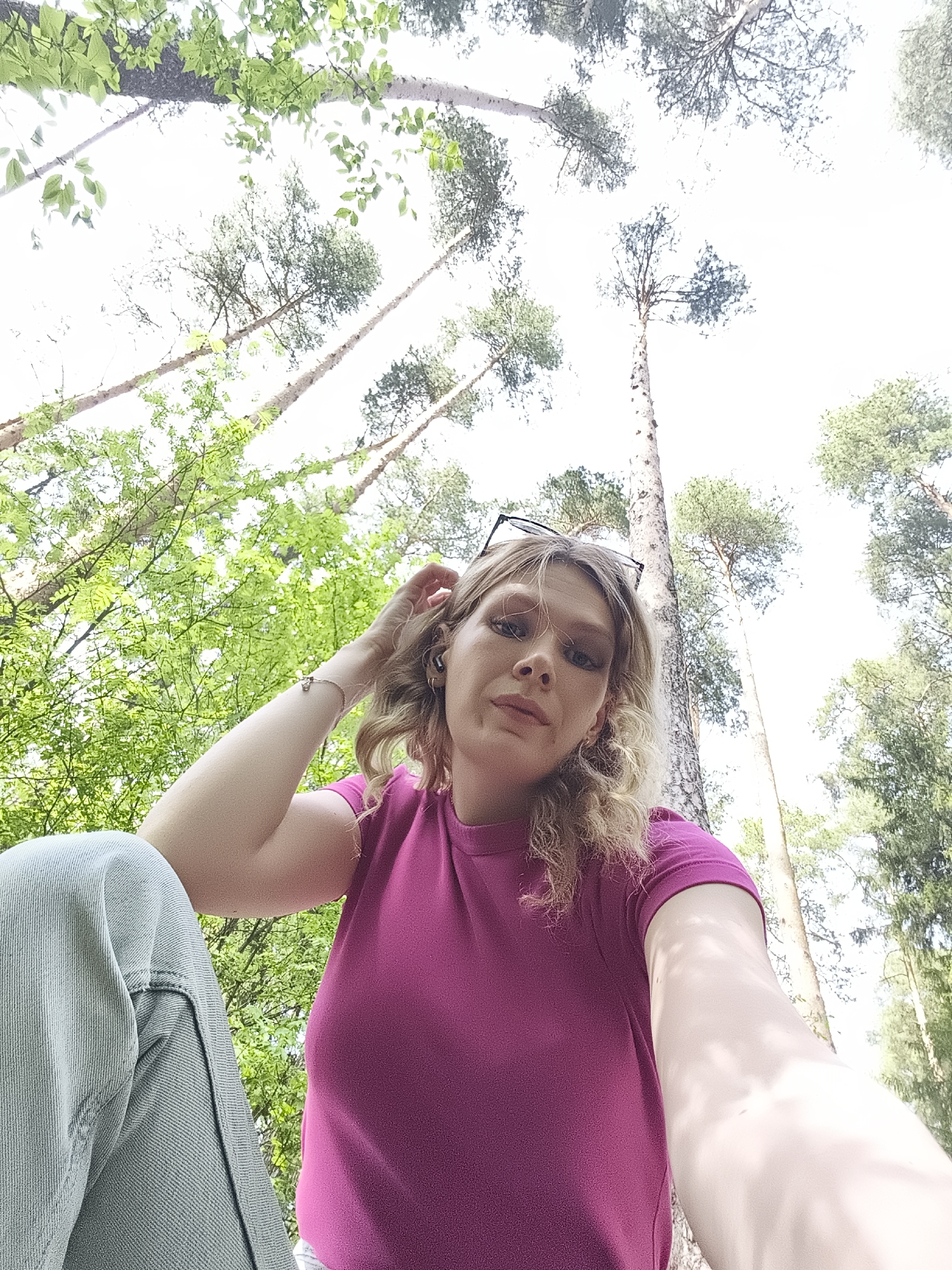 A person is angled upward, looking toward the camera, in a wooded area. The person wears a purple shirt and light-colored pants. Their blonde, curly hair is partially pulled back. Glasses rest on top of their head. Tall trees surround the person. Green leaves are visible. The location is presumably in Izhevsk, Russia.