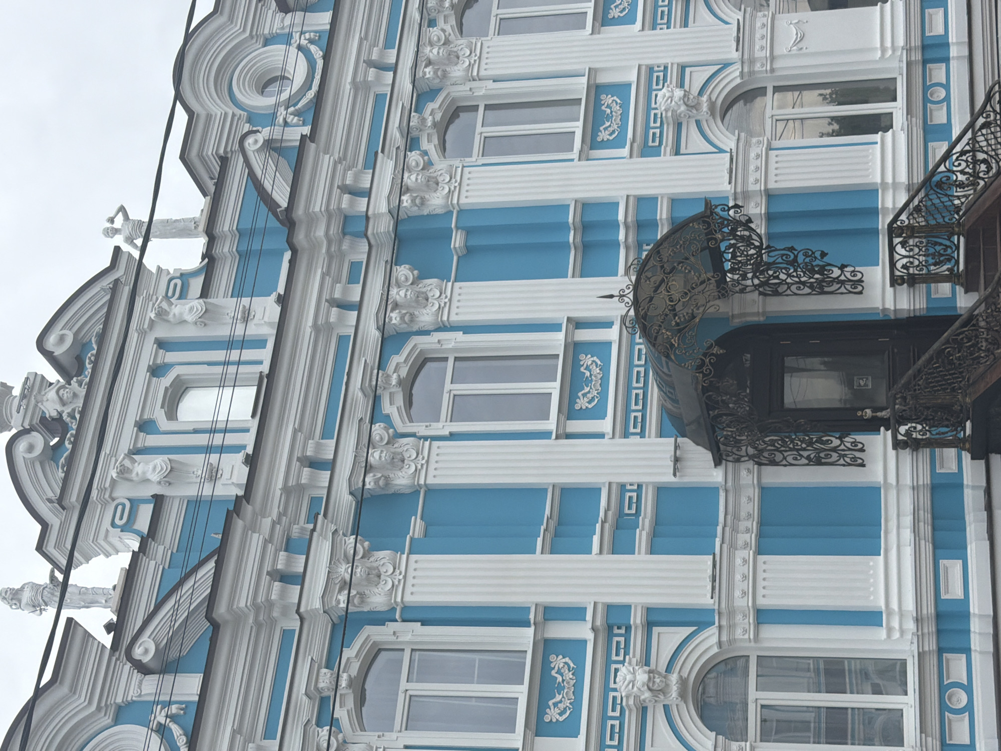 The image depicts a section of a building facade in Leningradskaya, Russia. The architectural style features a combination of white and blue colors, with ornate details such as sculpted figures along the roofline and decorative elements around the windows. Windows are visible on multiple floors, and a wrought-iron balcony with intricate designs projects from the building's exterior. The sky is overcast, suggesting a cloudy day.