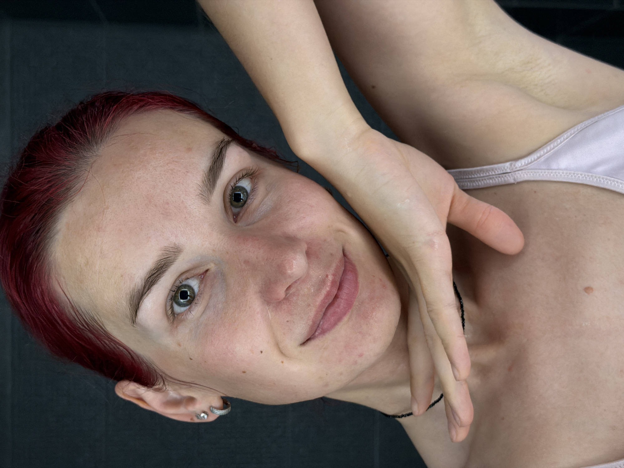 A close-up, front-facing photograph captures a young woman with bright red hair against a dark gray, textured background. She is looking directly at the camera, her head tilted slightly. Her left arm is bent, with her hand resting gently on her chin and neck. A delicate black beaded necklace is visible around her neck. She is wearing a pale lavender-colored tank top with thin straps. The lighting is soft, highlighting the natural texture of her skin, including some light blemishes and freckles. The image appears to be taken indoors. There is no indication of the location being in Chaikovsky, Russia.