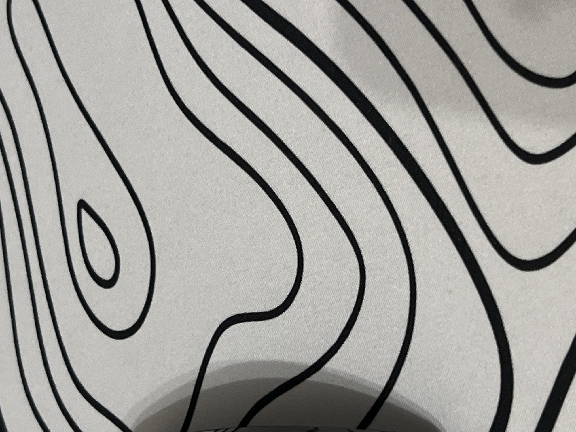 The image displays a close-up, top-down view of a white fabric with a pattern of thick, black, wavy lines. The lines vary in thickness and curvature, creating a sense of organic flow across the textured surface. The lighting suggests a soft, ambient source, casting subtle shadows that emphasize the fabric's weave and the depth of the pattern. There are no discernible landmarks or environmental cues to suggest a specific location, nor are there any people, subjects, or actions depicted. The image is purely a photographic study of a textile design.