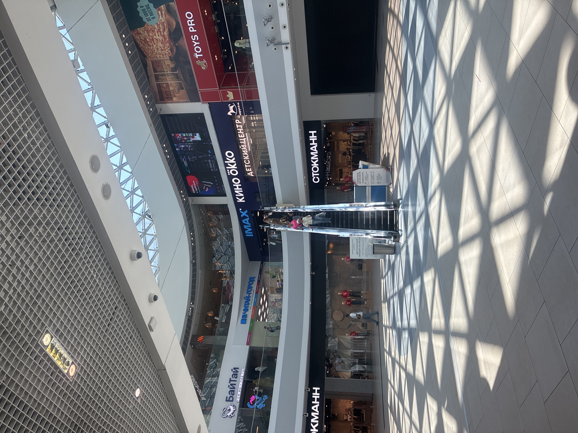 The image shows an interior view of a modern shopping mall, possibly in Sochi, Russia, with natural light streaming in from a large, geometric skylight. The architecture features multiple levels, escalators, and various store facades. Prominent store signs include "БайТай" (BaiTai), "Читай-город" (Chitay-gorod), "IMAX", "КИНО ÖKKÖ" (Kino ÖKKÖ), "ДЕТСКИЙ ЦЕНТР" (Detskiy Tsentr - Children's Center), "Toys PRO", and "СТОКМАНН" (STOKMANN). Several people are visible, including shoppers on escalators and walking on the floor. The floor is tiled and reflects the geometric patterns of the sunlight, creating a dappled effect. Security cameras are mounted on the ceiling. The overall impression is of a bustling commercial space during daylight hours.