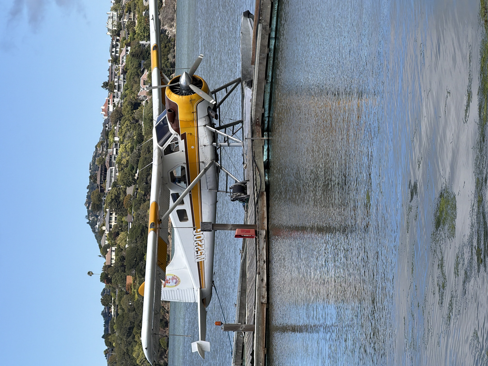 A white and yellow floatplane, identified by the registration number N5220G, is docked at a wooden pier on a body of water. The airplane has a single engine and a propeller. The background features a lush green hillside dotted with houses. The sky is clear and blue. The scene is likely located in Marin City, United States of America, given the distinctive architecture and landscaping often found in the region. There are no people visible in the image. The pier is weathered and shows signs of use, with a red fire extinguisher and a coiled rope visible on its surface.