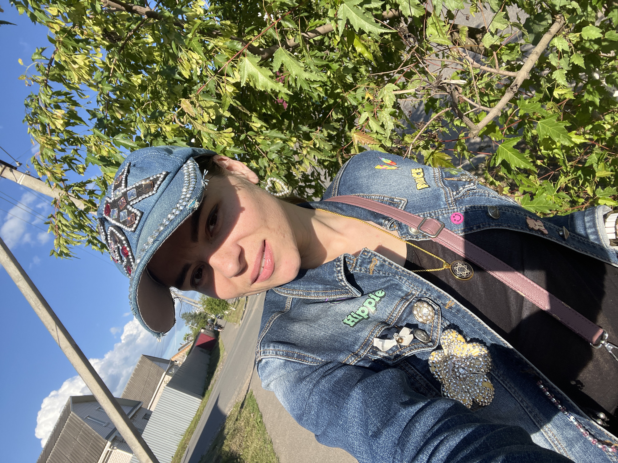 This is a selfie taken outdoors by a person wearing a denim jacket and a denim baseball cap. The person is smiling and looking towards the camera, with sunlight casting a shadow on their face. The background shows green trees, a clear blue sky with some clouds, a utility pole, and houses with red roofs, consistent with a rural or suburban setting. The denim jacket is decorated with various patches and pins, including one that says "Hippie" and a sparkling flower brooch. The person is also wearing a necklace with a Star of David pendant and has a pink purse strap visible. The time of day appears to be daytime, with bright sunlight. The overall mood seems cheerful and relaxed. The location context provided is Krasnoe-na-Volge, Russia.