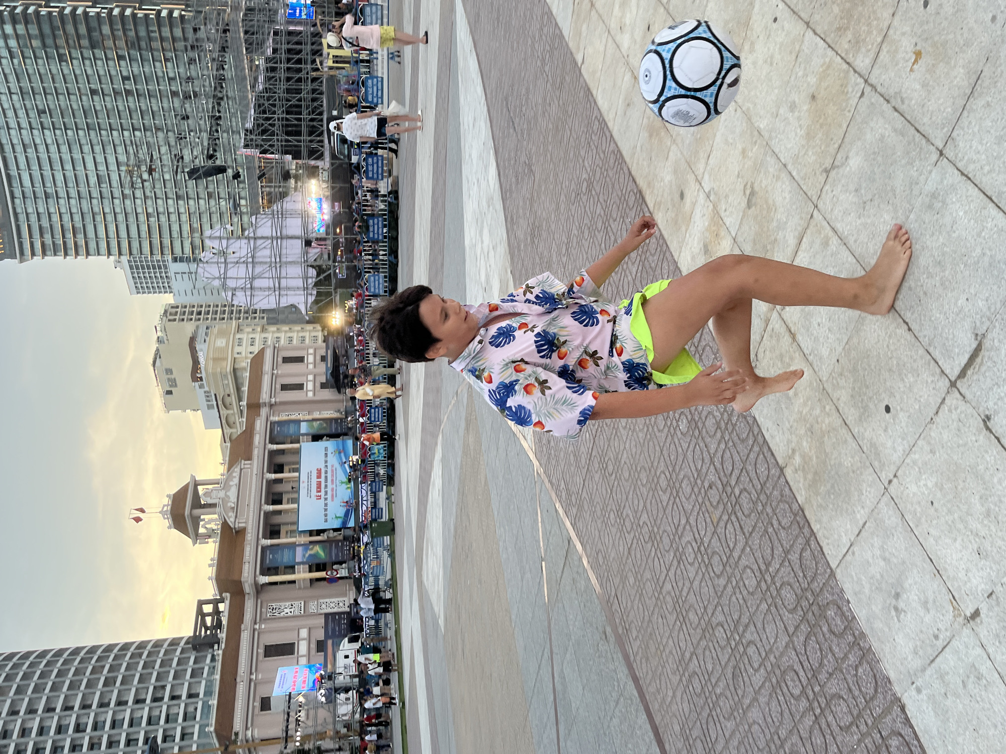 An athletic young boy, wearing a tropical-print short-sleeved shirt and neon green shorts, is barefoot and actively playing with a black and white soccer ball in a wide, paved plaza. The boy is captured mid-action, with his right leg extended as if kicking the ball, which is in the air to his right. His gaze is focused on the ball.
Behind him, the plaza is vast and features a patterned paving stone surface. In the background, a large, ornate building with a brown roof and white columns stands prominently, topped with two red flags. This building appears to be a governmental or cultural center. Further in the background, modern high-rise buildings dominate the skyline, suggesting an urban environment. There are also stages and scaffolding set up, indicating an event or festival is either in progress or about to begin. Several people are visible in the distance, some walking, others gathered, suggesting a public gathering. The sky above is a pale, hazy blue with hints of yellow, indicative of either early morning or late afternoon.
The location is likely the 2/4 Square (Quảng trường 2 tháng 4) in Nha Trang, Vietnam, a well-known central plaza often used for public events and gatherings, which is situated near prominent buildings and the coastline.