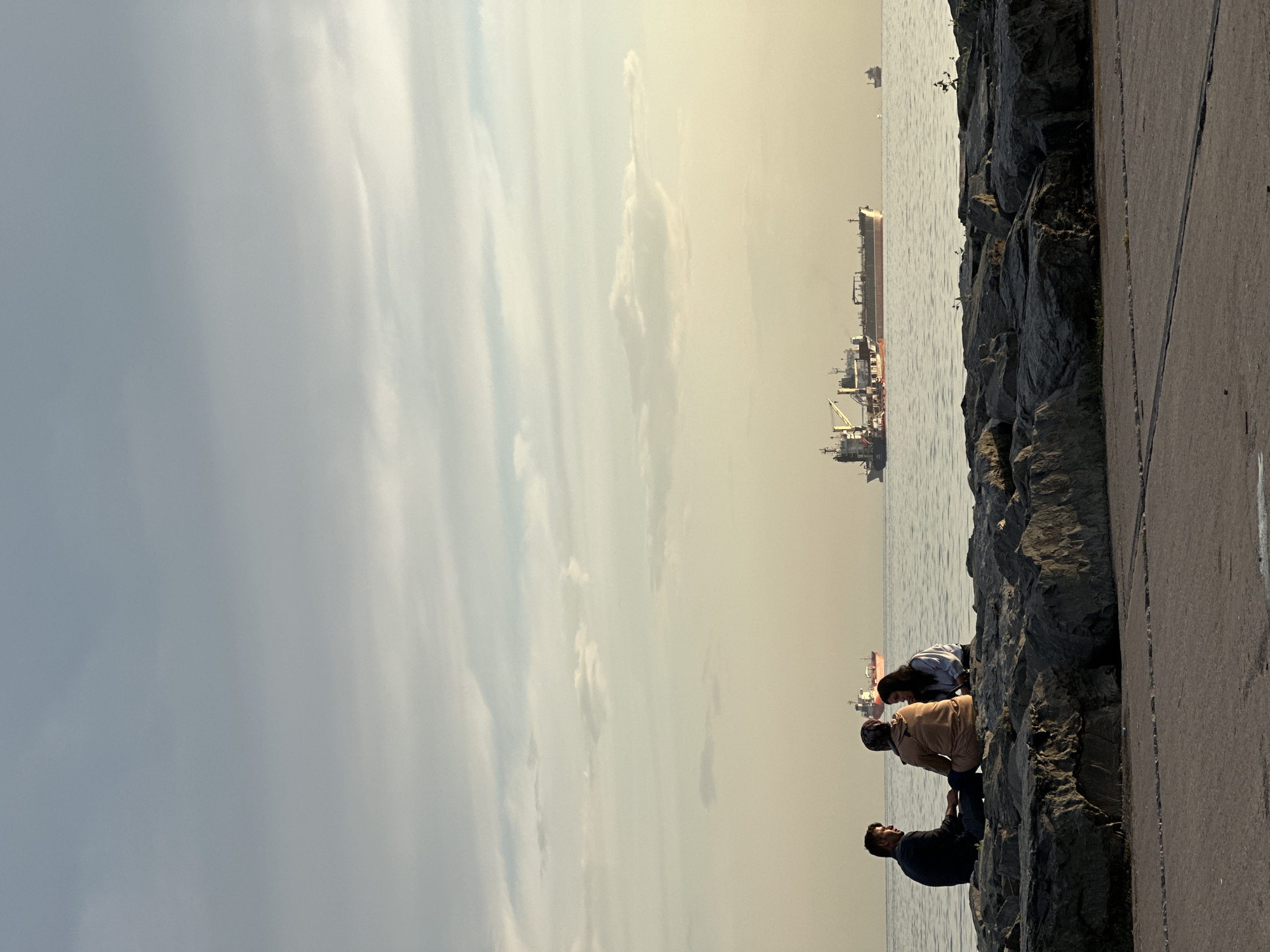 In Bakırköy, Turkey, three individuals are seated on a rocky shoreline. A man is on the left, and two women are to his right. They appear to be engaged in conversation. In the background, several large cargo ships are visible on the water. The sky is overcast with soft, diffused light. The foreground shows a paved walkway and large stones, suggesting a public waterfront area.