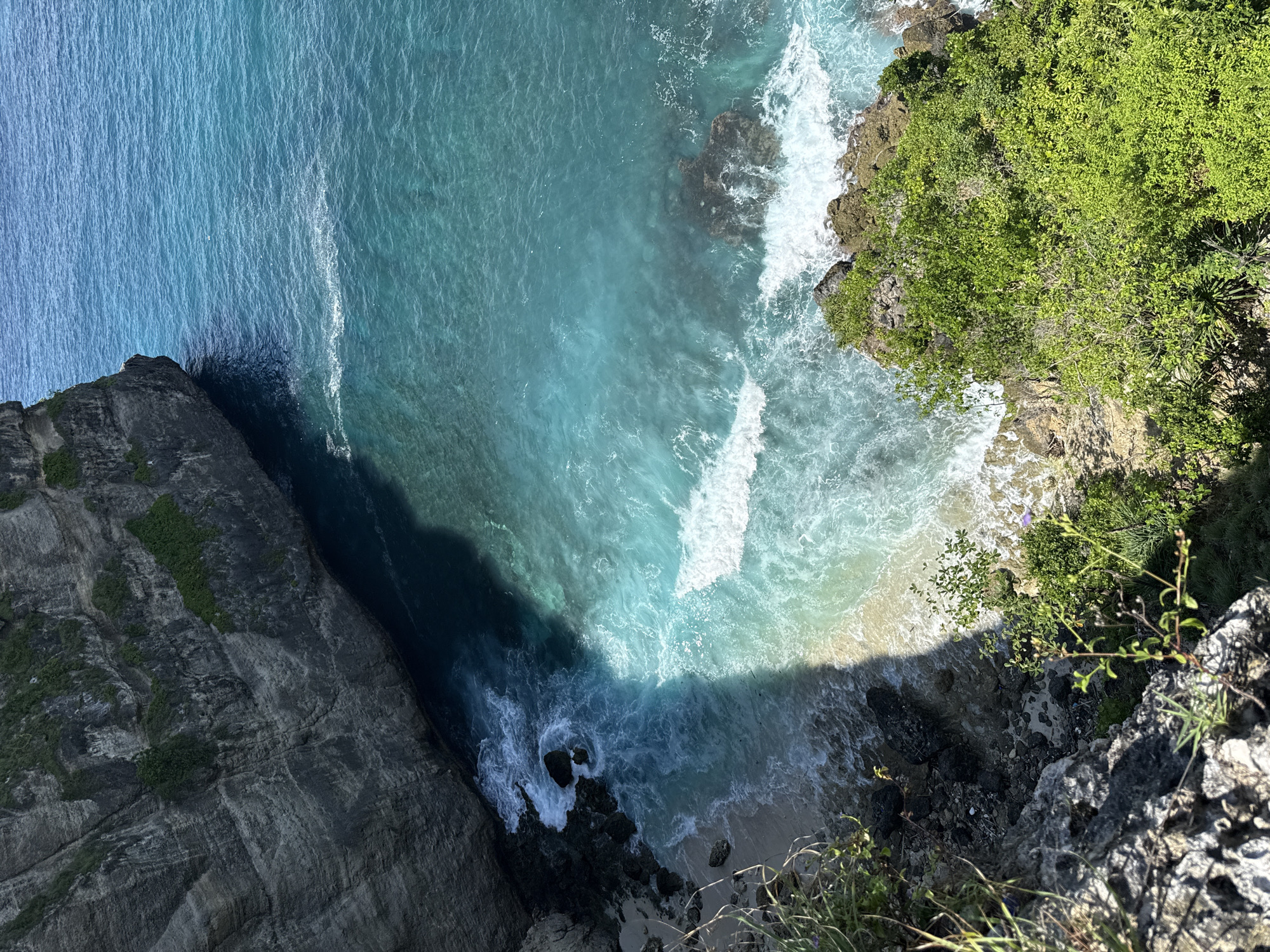 A high-angle shot depicts a coastal scene in Pejukutan, Indonesia. Dominating the left and right sides are rugged cliffs, with the left one appearing darker and more textured, showing various geological layers. The right cliff is covered with dense green vegetation. The water between the cliffs showcases varying shades of blue, from a lighter turquoise to a deep blue, influenced by sunlight and depth. White waves crash against the shore and the base of the cliffs, indicating active water movement. A small sandy beach is visible at the bottom center. The perspective suggests the shot was taken from a high vantage point, likely overlooking the coastline. No people are visible in the scene.
