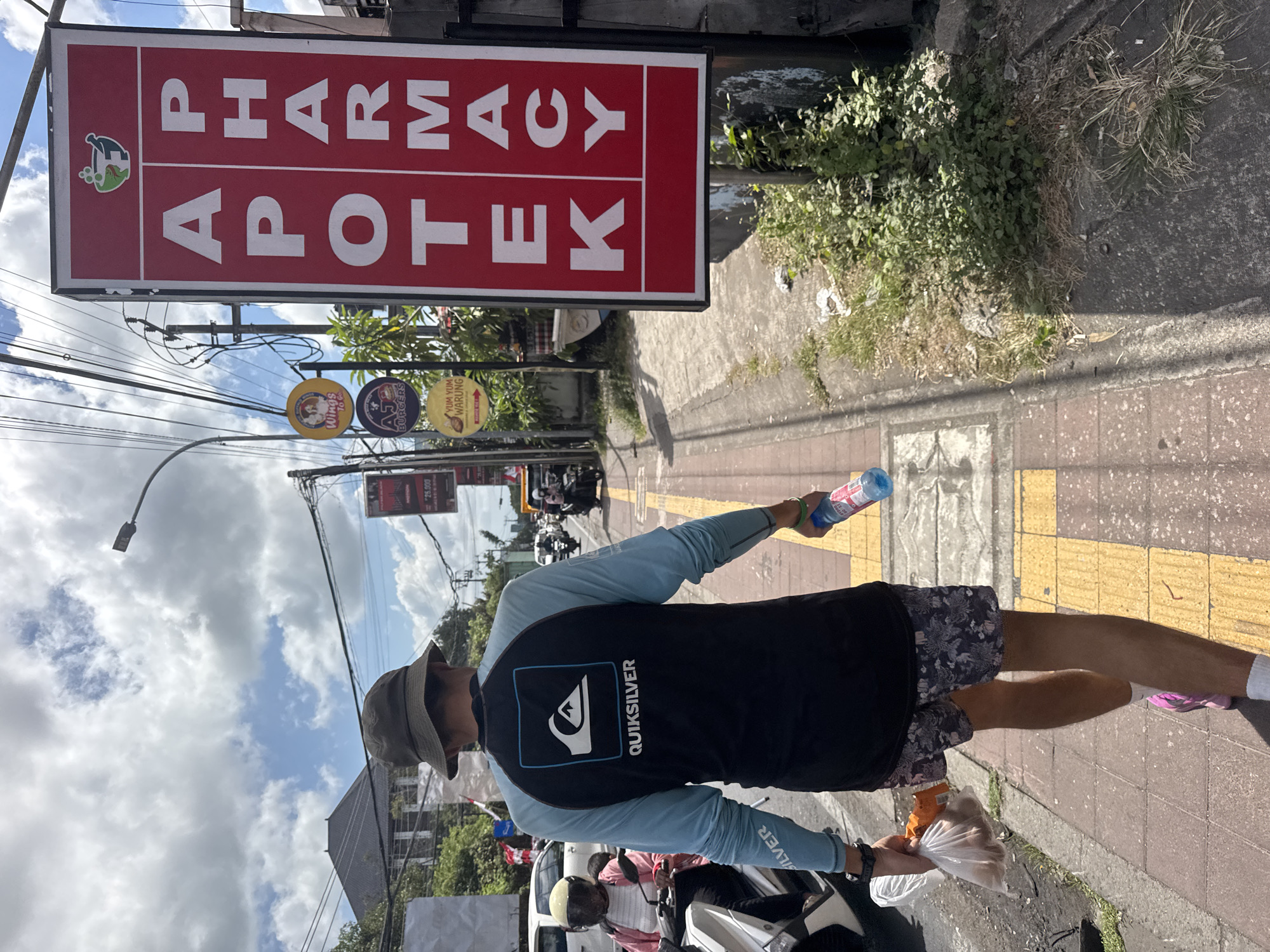 The image depicts a street scene in Kuta Selatan, Indonesia, with a person walking on a sidewalk. The person, viewed from behind, is wearing a bucket hat, a long-sleeved Quiksilver rashguard in blue and black, and patterned shorts. They are carrying a plastic bag with food items in their left hand and a blue water bottle in their right. The sidewalk is paved with red bricks and features yellow tactile paving. To the right of the sidewalk is a prominent red sign with white lettering that reads "APOTEK PHARMACY," indicating a pharmacy. Several circular signs for food establishments like "Wings To Go," "AJ Burgers," and "Yum Yum Warung" are visible on poles in the background. The environment includes a street with vehicles, including motorcycles, and overhead power lines. The sky is partly cloudy with a bright sun, suggesting daytime. The scene conveys a sense of everyday life on a street in a tropical urban setting.