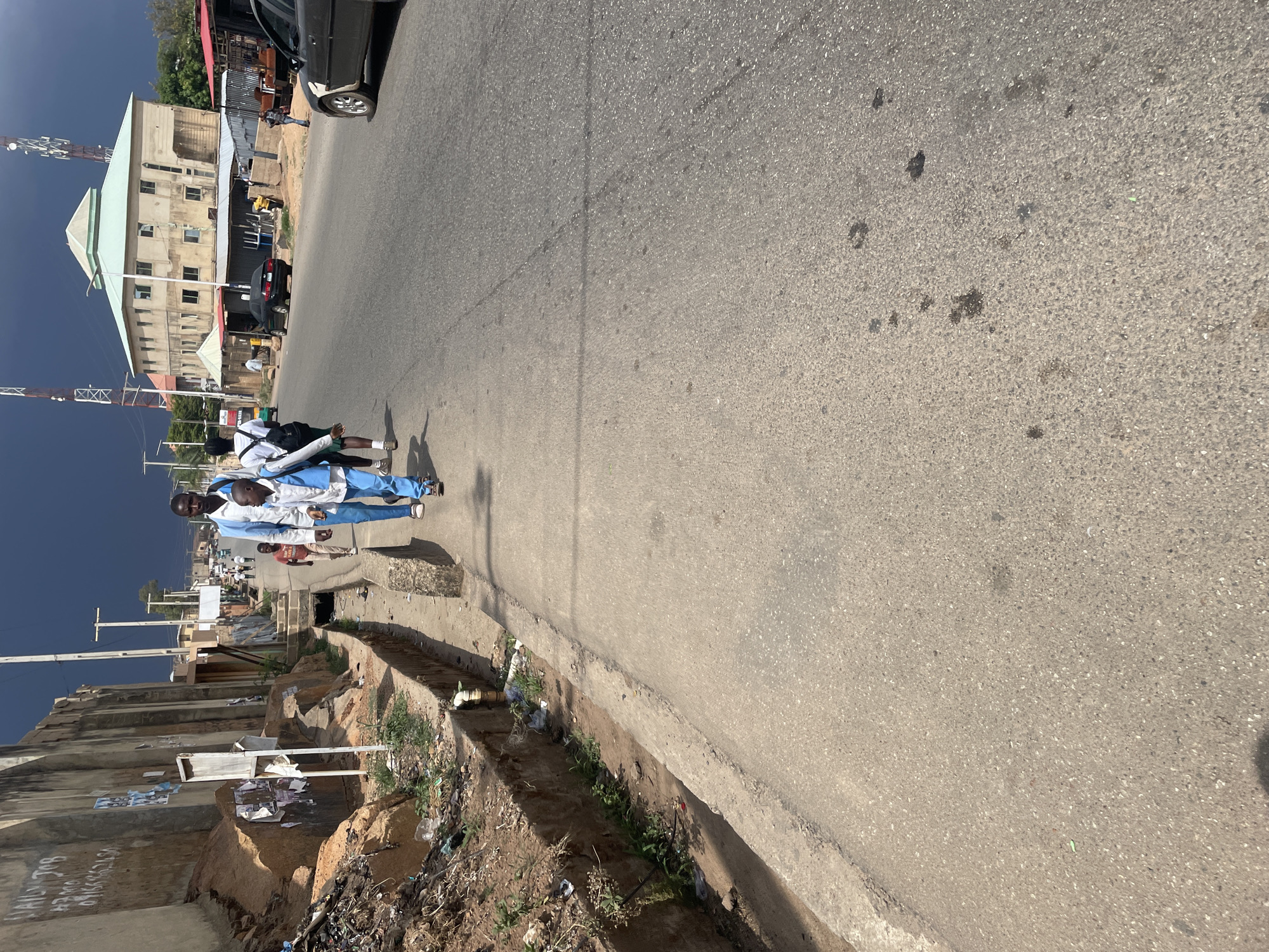 The image shows a street scene in Jos, Nigeria. Two males walk along the edge of a paved road. The males are wearing light blue and white outfits. The road is adjacent to a concrete curb, with a drainage ditch on the other side. There is trash accumulated in the ditch. Buildings are visible on the left side of the road. A black car is parked on the right side of the road. The sky is dark.