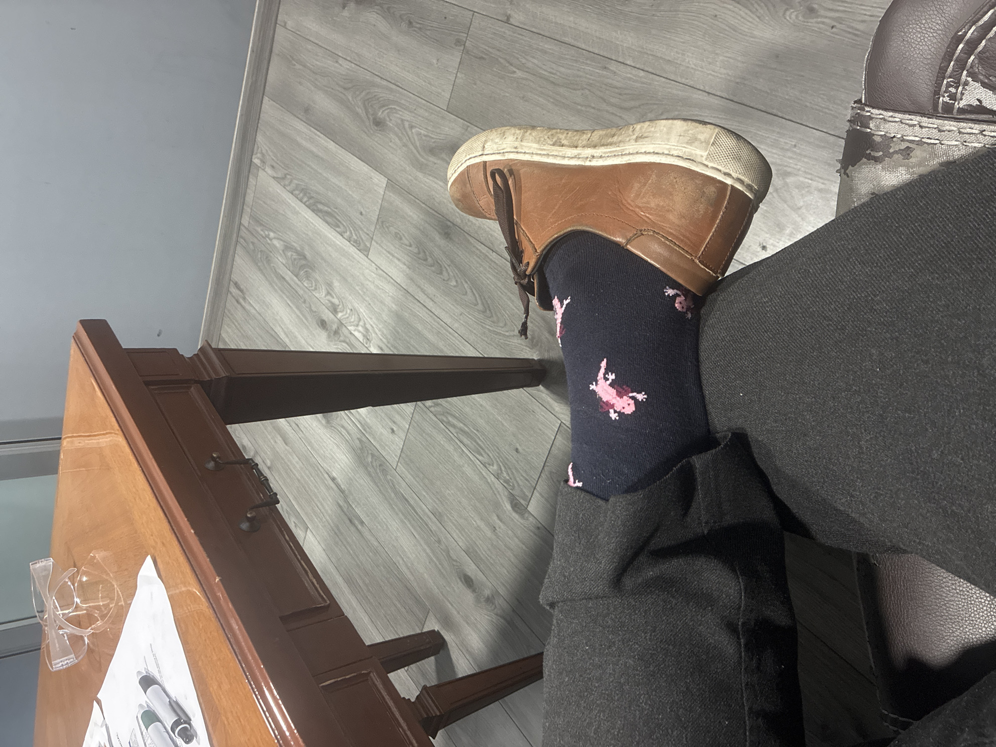 A person wearing black jeans, a brown leather shoe on their right foot, and a dark blue sock with pink flamingoes sits next to a brown wooden desk. The desk has a pair of clear plastic safety glasses and papers with markers on top. The flooring is grey wood-patterned vinyl. The location is Cuautitlán Izcalli, Mexico.