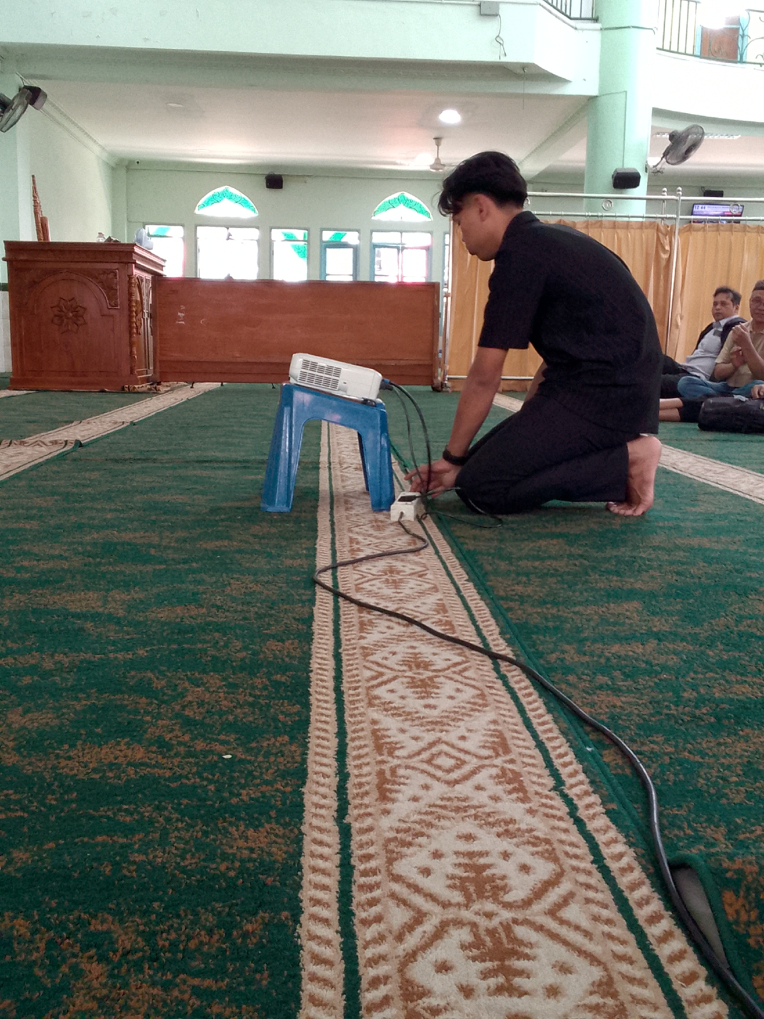A young man in a black shirt and pants kneels on a green carpet in a large room, possibly a mosque, given the presence of a wooden podium and arched windows. He is connecting cables to a white power strip on the floor, near a blue plastic stool supporting a white projector. In the background, several men are seated on the carpet, with one appearing to be speaking. The room has high ceilings, light-colored walls, and fans mounted on the walls. The flooring is carpeted with a green base and a patterned border. The overall scene suggests an event or gathering taking place indoors. The location is identified as Cimahi, Indonesia.