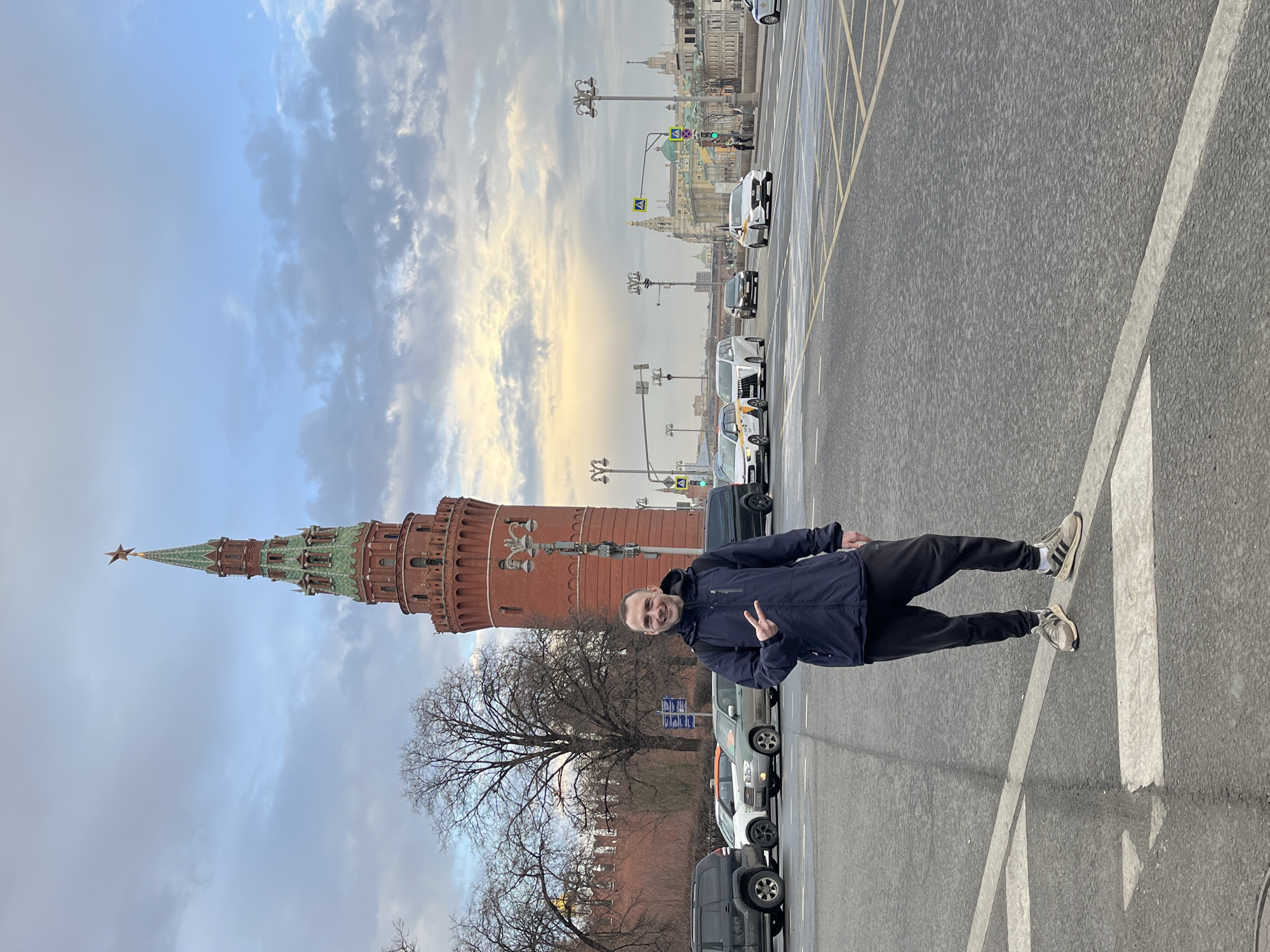 A man poses for a photo on a Moscow street in front of the Kremlin. The subject, a Caucasian man, stands facing the camera and gives a peace sign with his right hand. He wears a dark blue jacket, dark pants, and athletic shoes. In the background is the red brick Kremlin wall and the Spasskaya Tower, topped with a star. The street appears to be wet, with multiple parked cars and taxis visible. The sky is overcast with varied cloud formations.